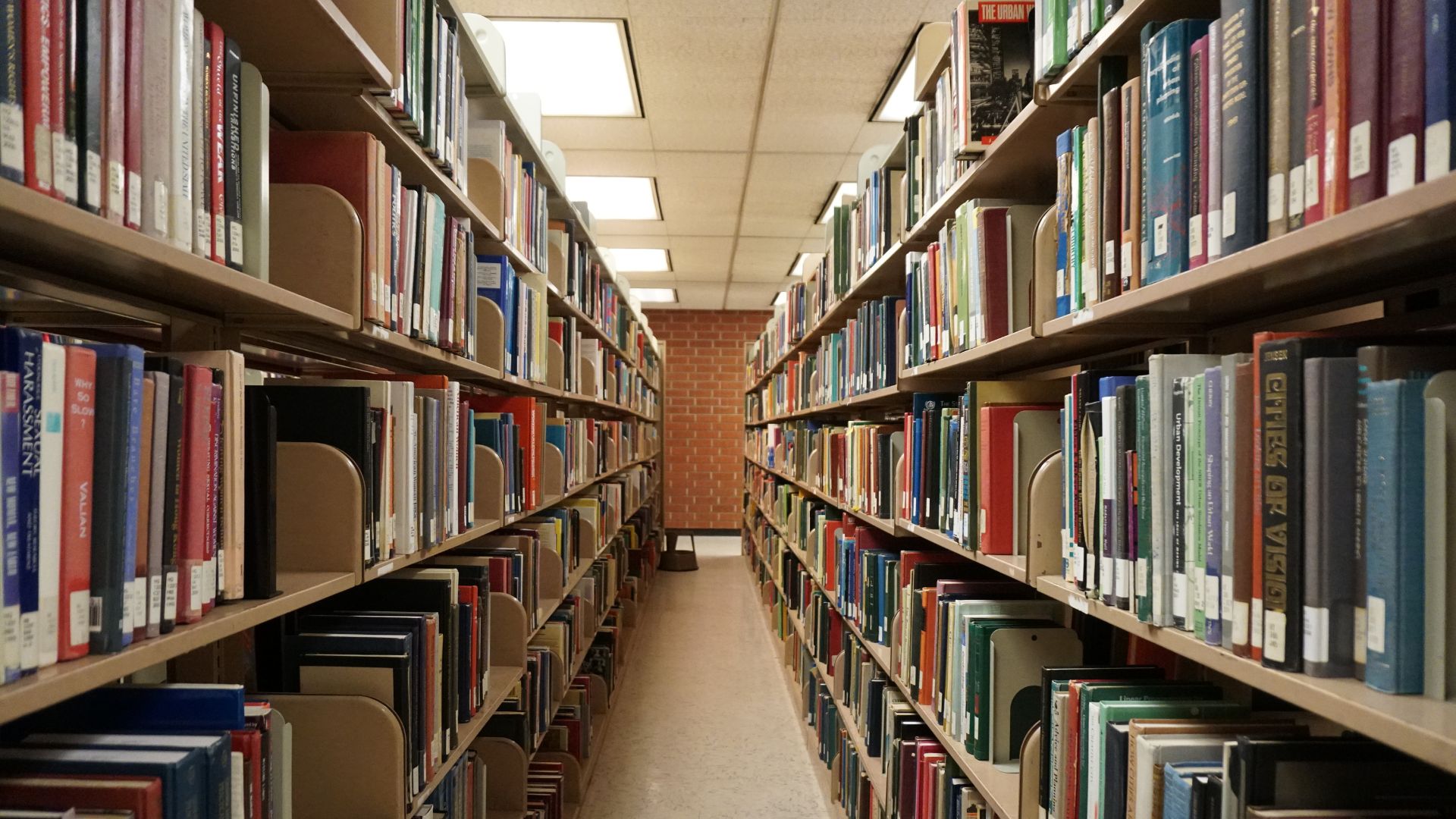A long, narrow aisle in a library lined with tall wooden bookshelves filled with various books on both sides, leading towards a brighter area at the far end.