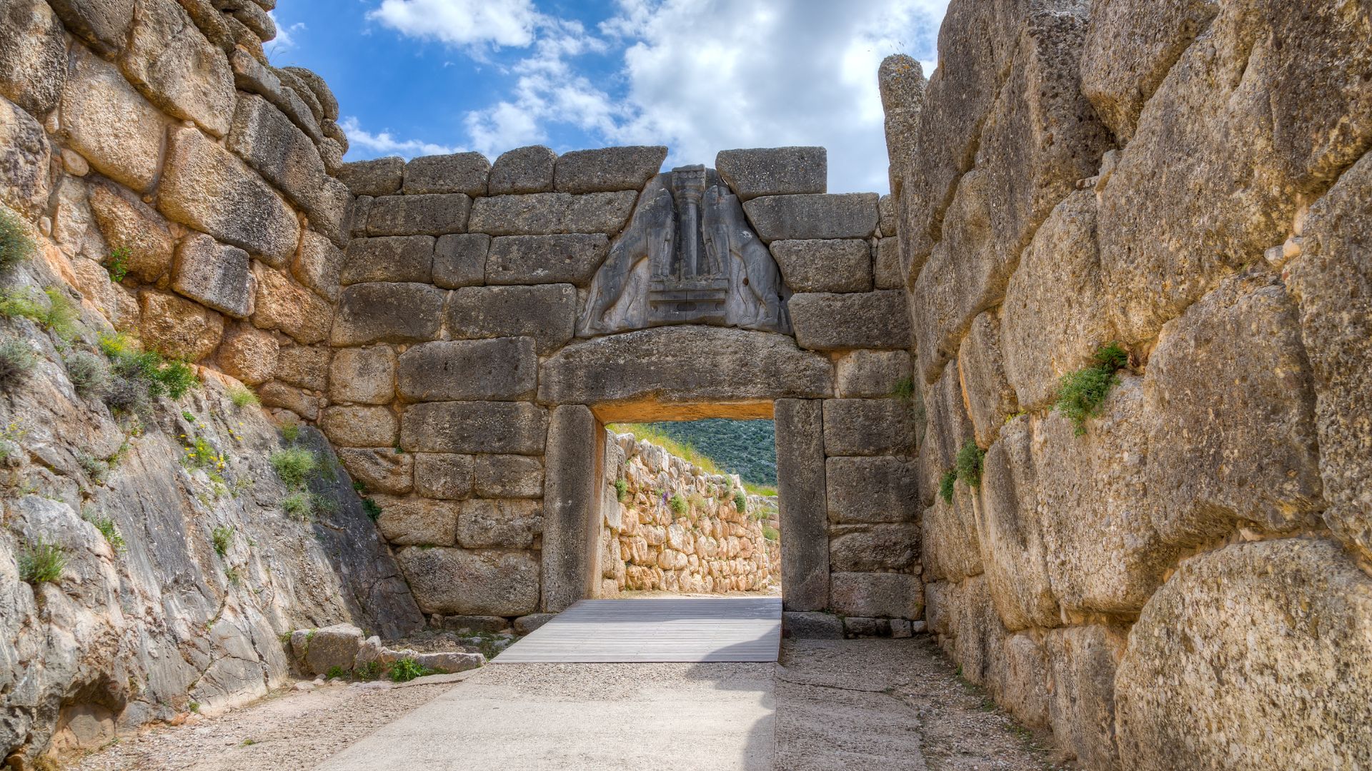 A view looking through the iconic Lion Gate at Mycenae, a massive stone entranceway with a prominent relief sculpture of two lionesses above the lintel, flanked by large cyclopean walls on either side under a partly cloudy sky.