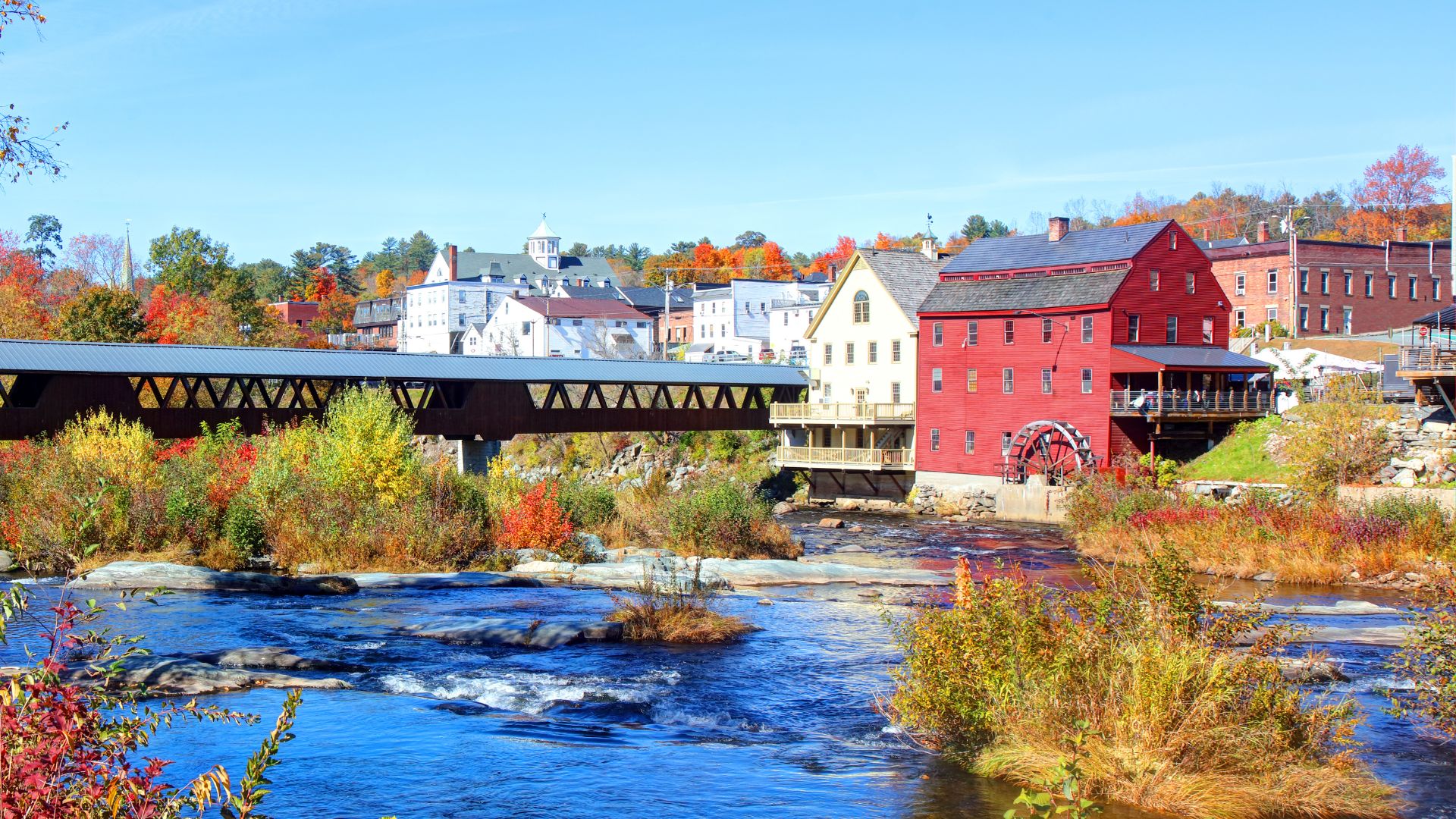 A vibrant fall scene in Littleton, New Hampshire, featuring a covered bridge spanning a river, with colorful autumn foliage and historic buildings, including a red mill with a waterwheel, lining the riverbank under a clear blue sky.