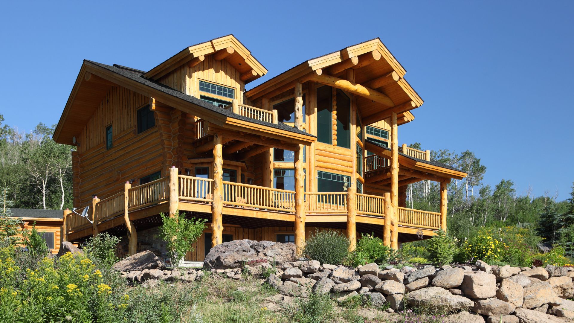 A large, modern log cabin with multiple levels and expansive decks, constructed from light-colored logs, sits on a rocky, landscaped incline under a clear blue sky.