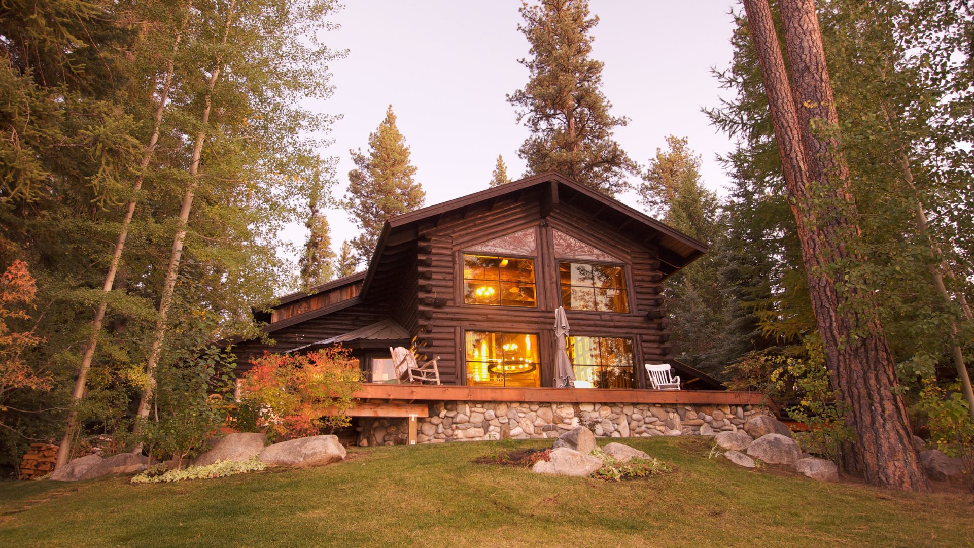 A rustic log cabin nestled among tall pine trees, featuring large windows that glow with warm light from within, and a spacious wooden deck with stone foundation.
