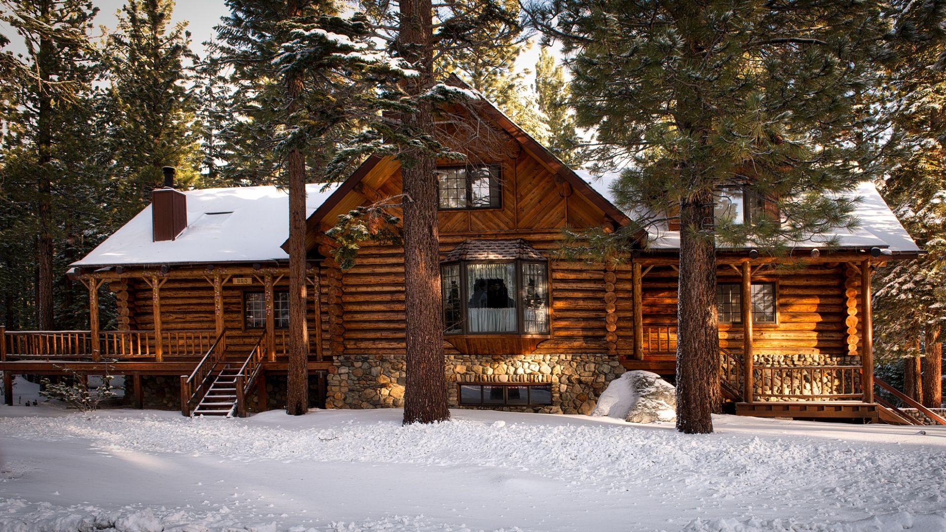 A rustic log cabin with a snow-covered roof and ground, nestled among tall pine trees in a winter setting.
