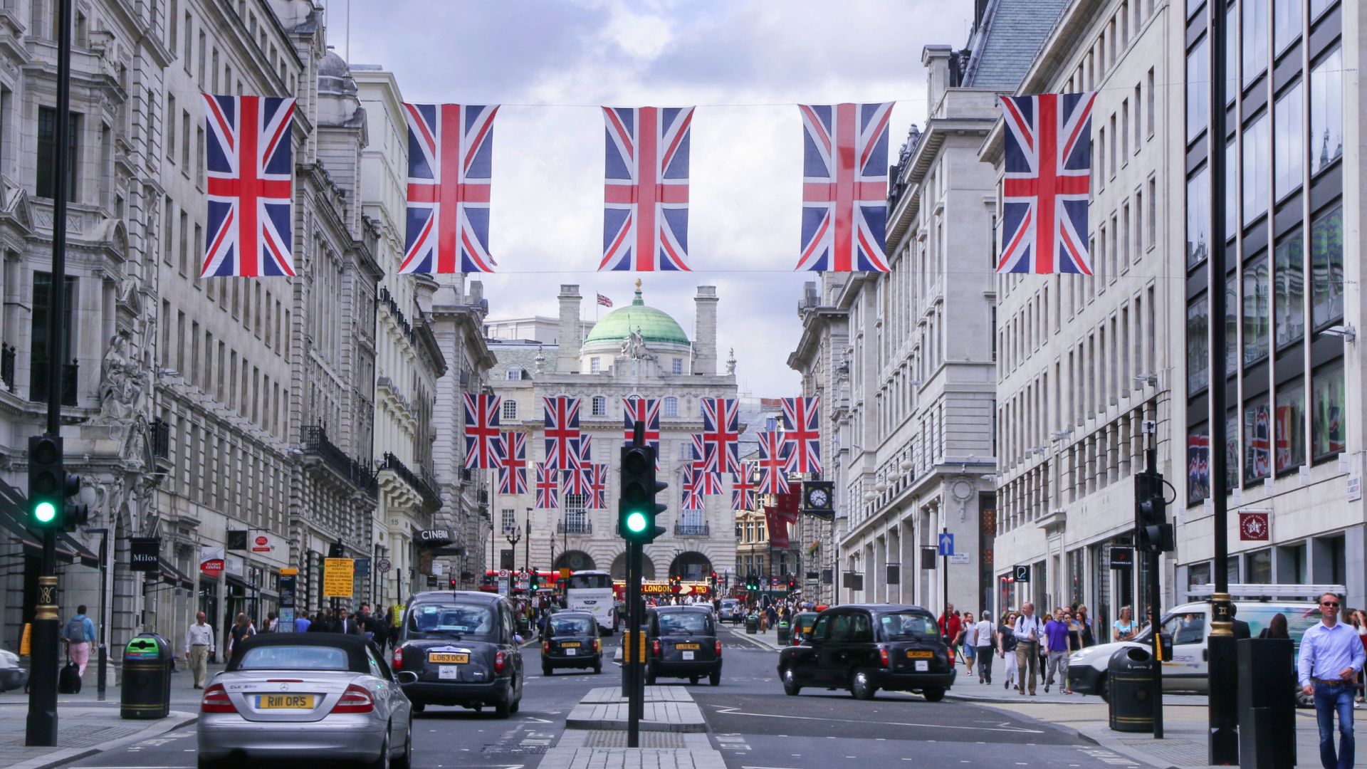 A bustling street in London, England, adorned with numerous Union Jack flags hanging overhead between buildings. The street features classic architecture, black taxis, and cars, with pedestrians on the sidewalks, all under a cloudy sky.