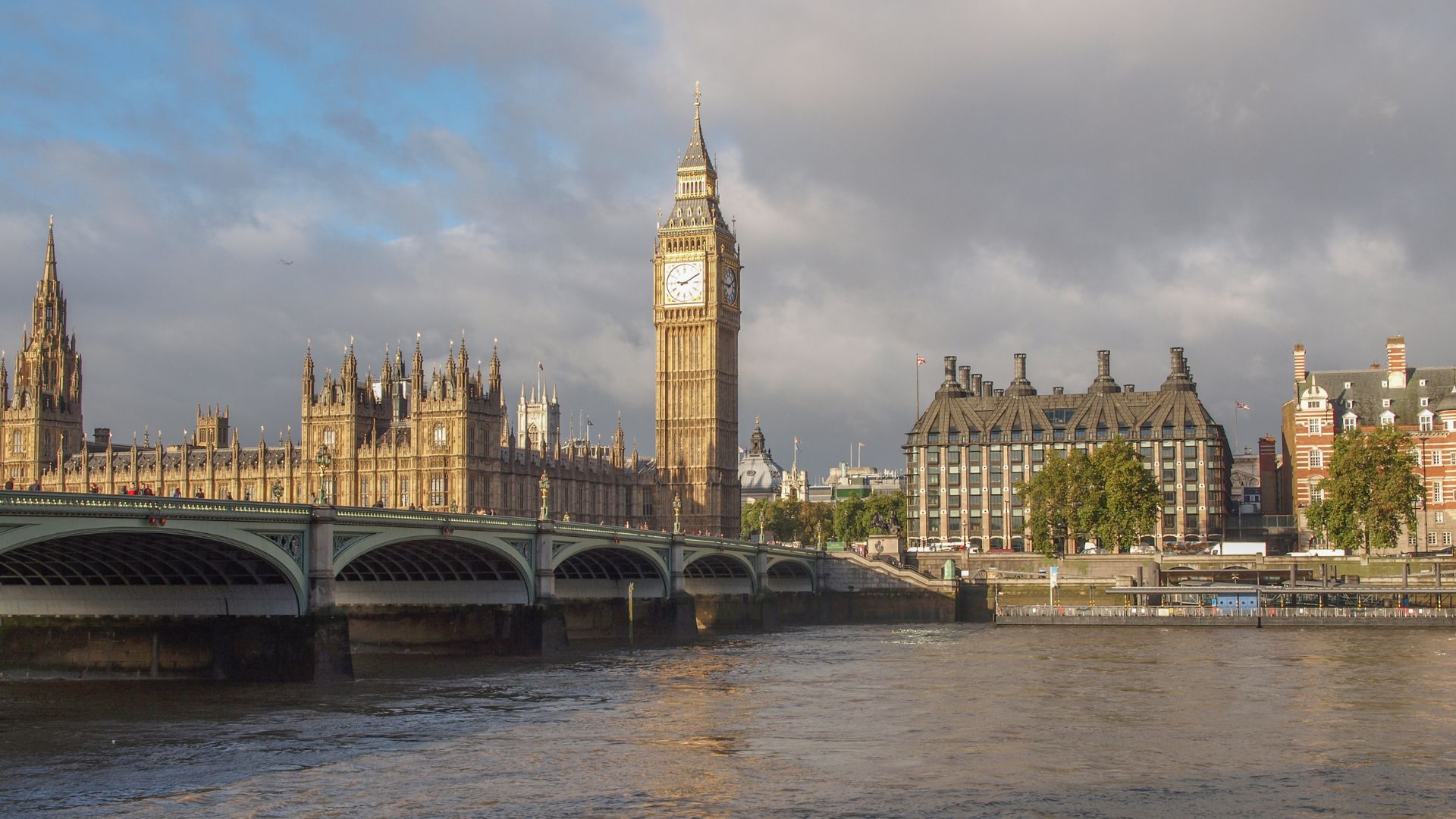 A wide shot of the Palace of Westminster and Big Ben in London, United Kingdom, seen from across the River Thames with Westminster Bridge spanning the river in the foreground under a partly cloudy sky.