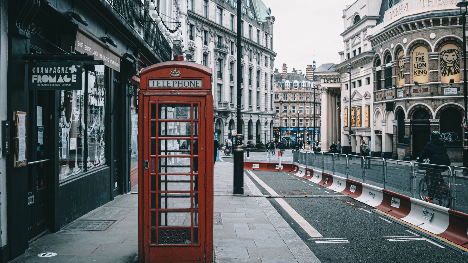 A classic red British telephone box stands prominently on a London sidewalk, with historic buildings and a street leading into the distance. The Lyceum Theatre, home to "The Lion King" musical, is visible in the background.