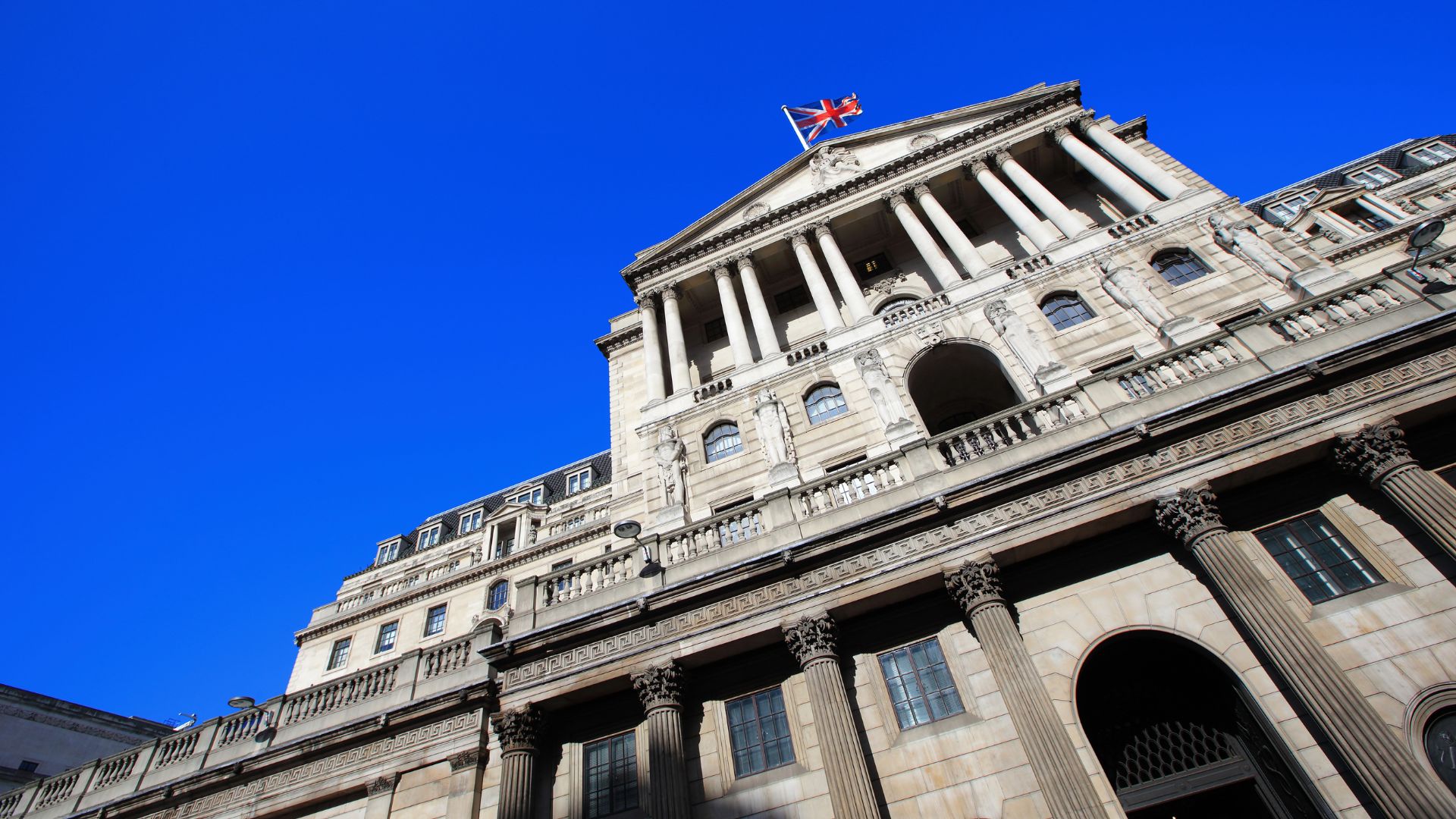A low-angle shot of the grand, classical facade of the Bank of England building in London, featuring columns and a British flag flying atop its roof, set against a vibrant blue sky.