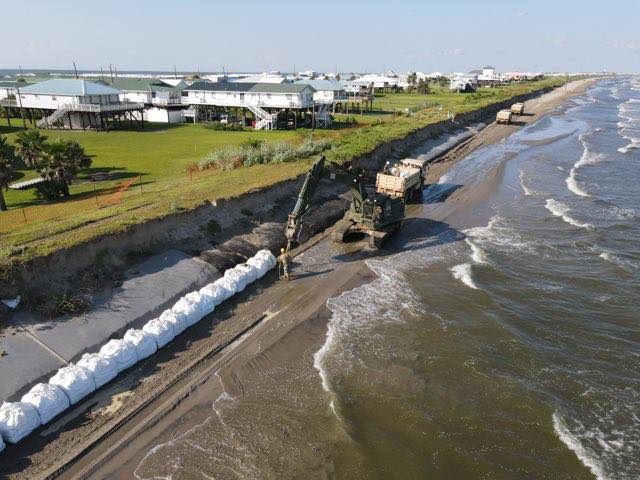 Louisiana Army National Guard soldiers and military vehicles reinforcing a levee with sandbags and equipment in preparation for Hurricane Laura, under cloudy skies with a sense of urgency.