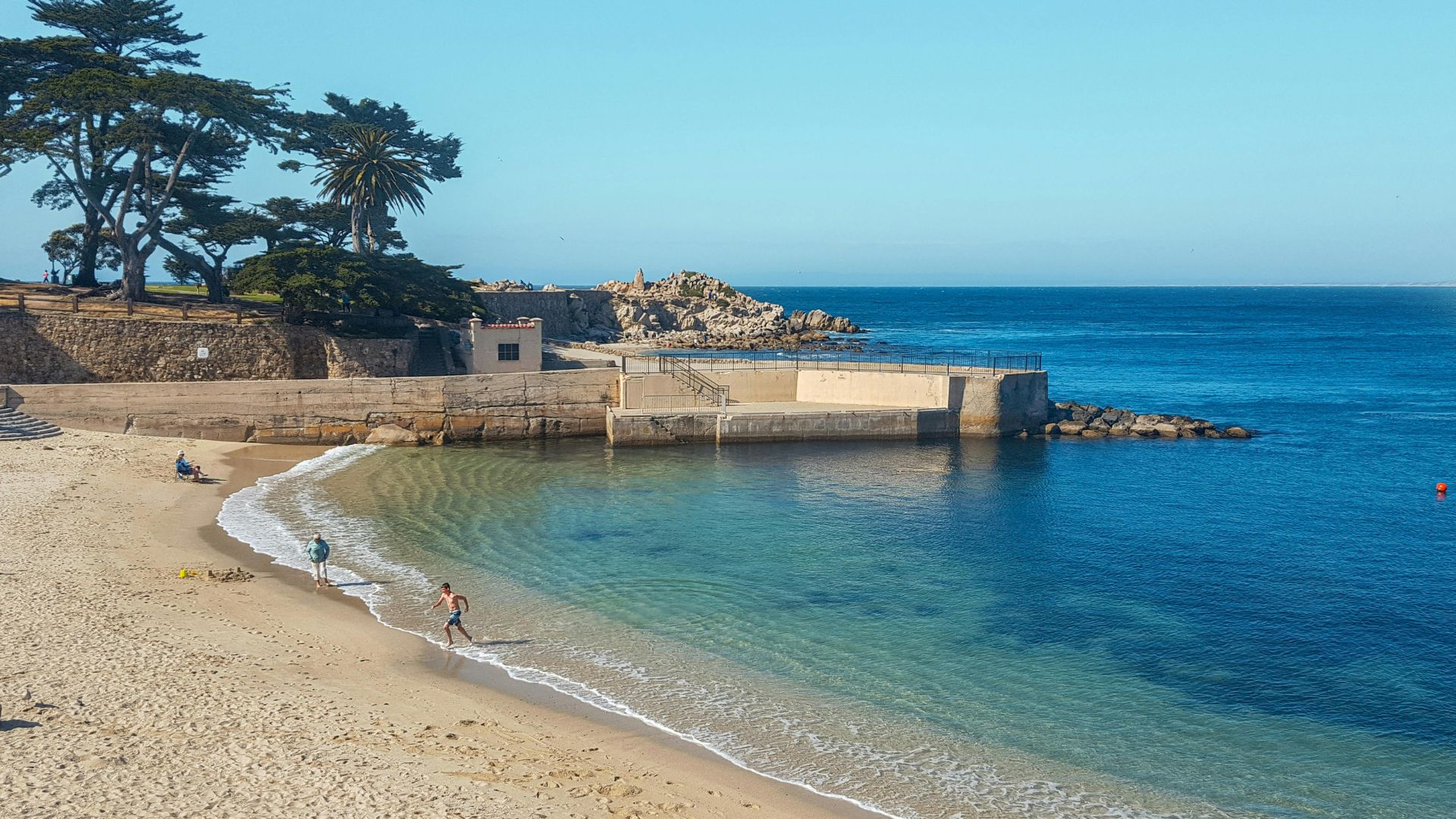 A sunny coastal scene featuring a sandy beach curving around a calm, clear turquoise bay with a concrete pier and rocky shoreline on the right. In the background, green trees and buildings are visible on a higher land area, and a person is walking on the beach near the water's edge.