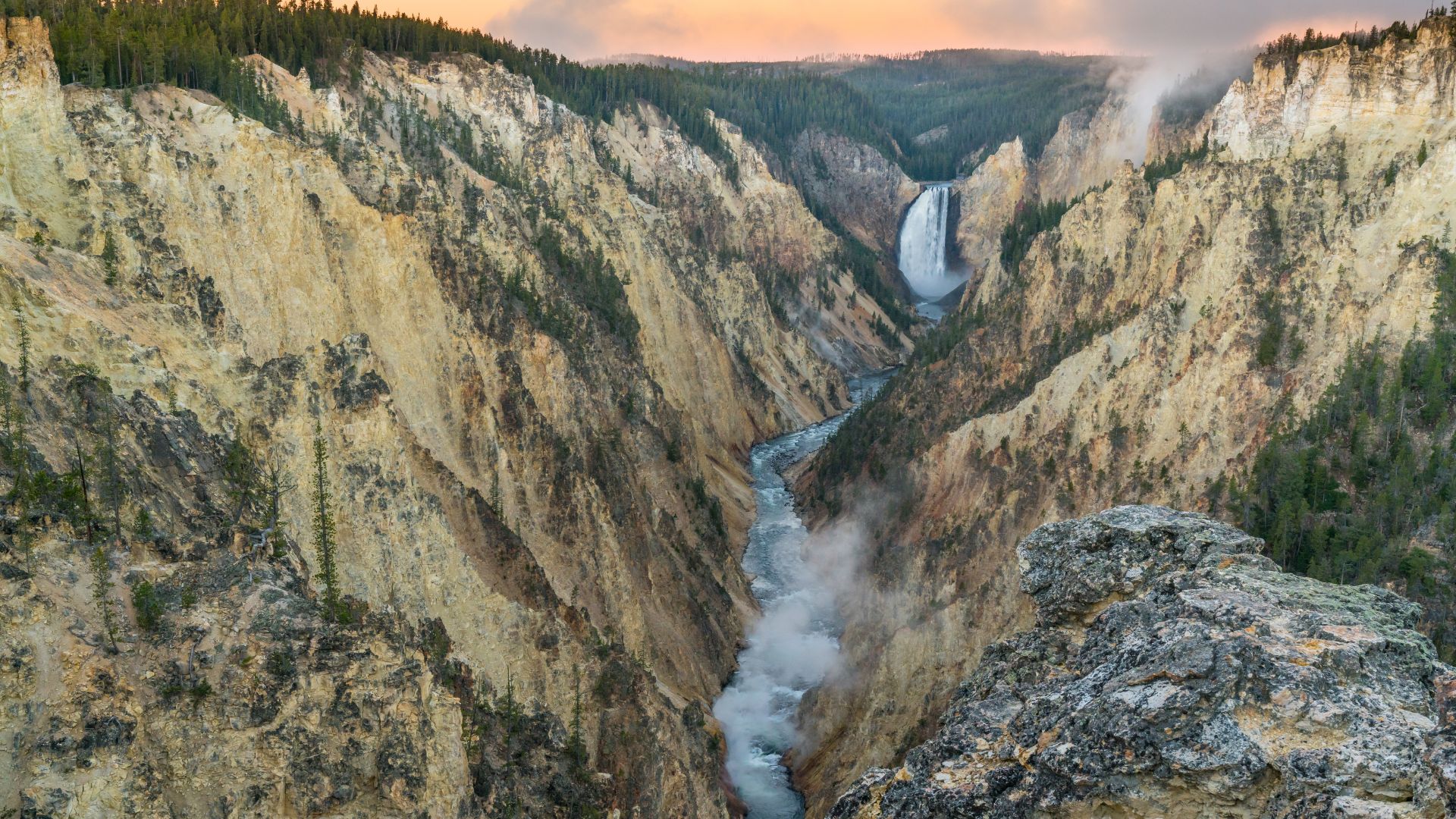 A majestic view of the Lower Yellowstone Falls plunging into the Grand Canyon of the Yellowstone, with a river winding through the rugged, tree-lined canyon walls under a dramatic sky.