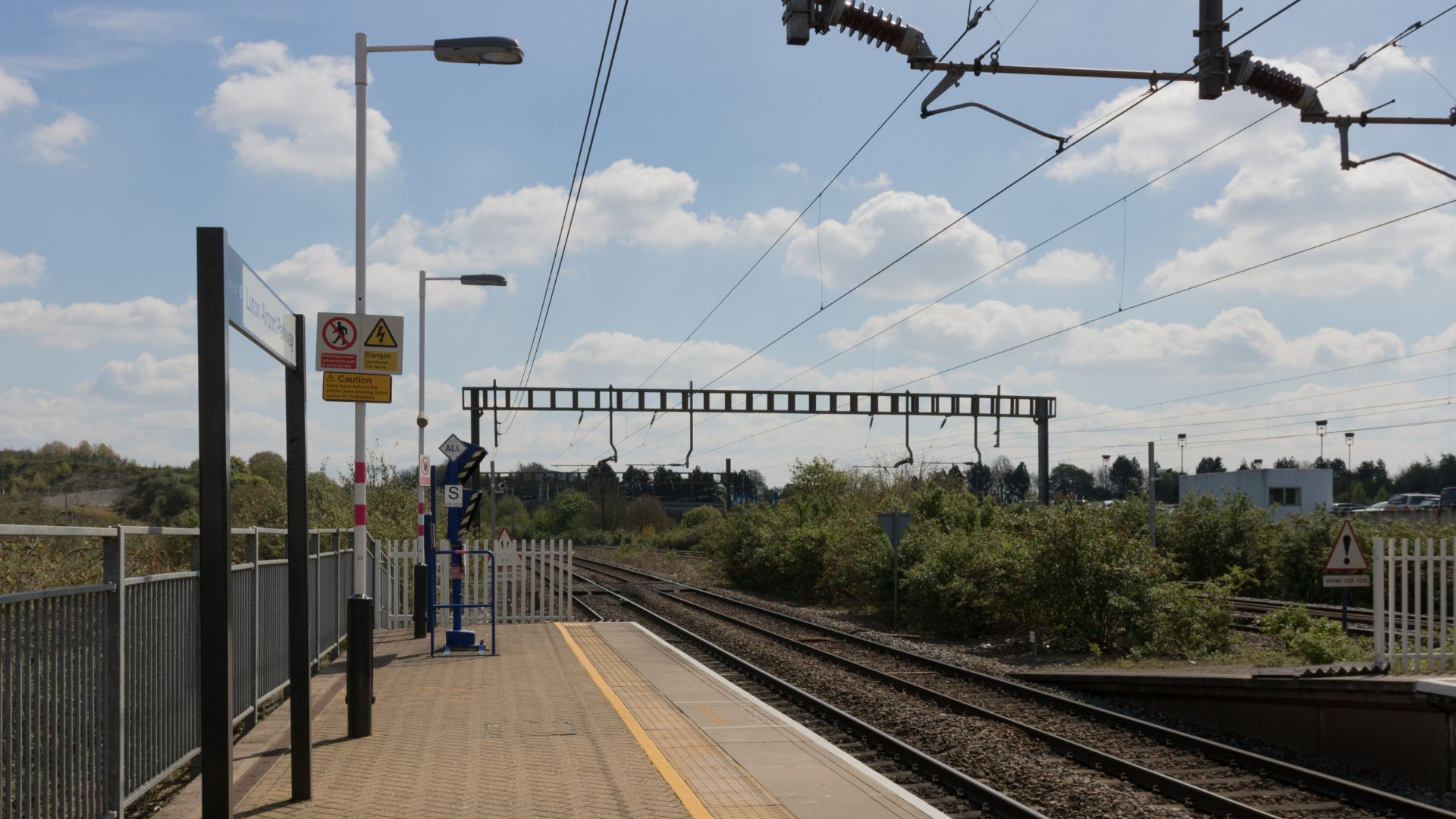 A bright, sunny image of a train station platform with tracks extending into the distance under overhead power lines, featuring a signal gantry, lampposts, and a "no entry" sign on the left.
