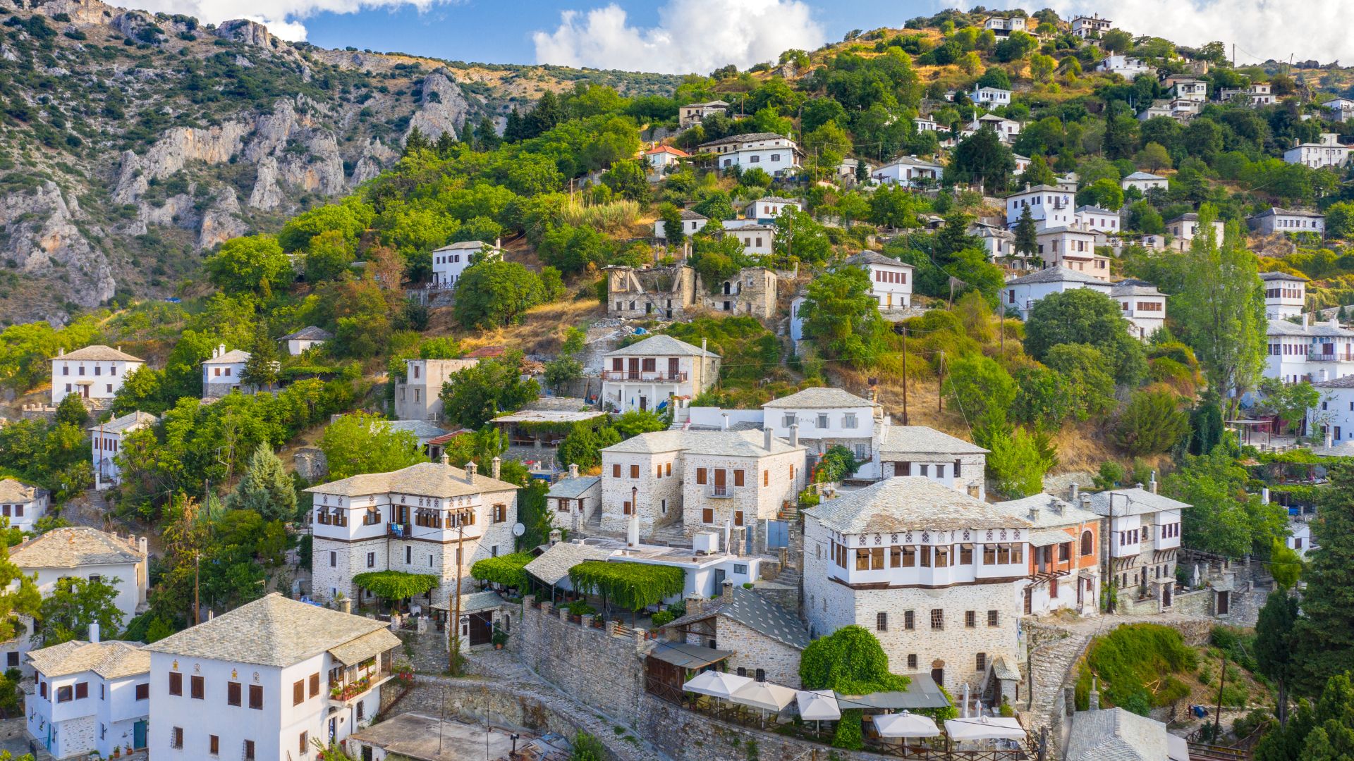 A hillside village in Greece, featuring traditional stone houses with white walls and red-tiled roofs nestled among lush green trees, overlooking a valley with distant views.