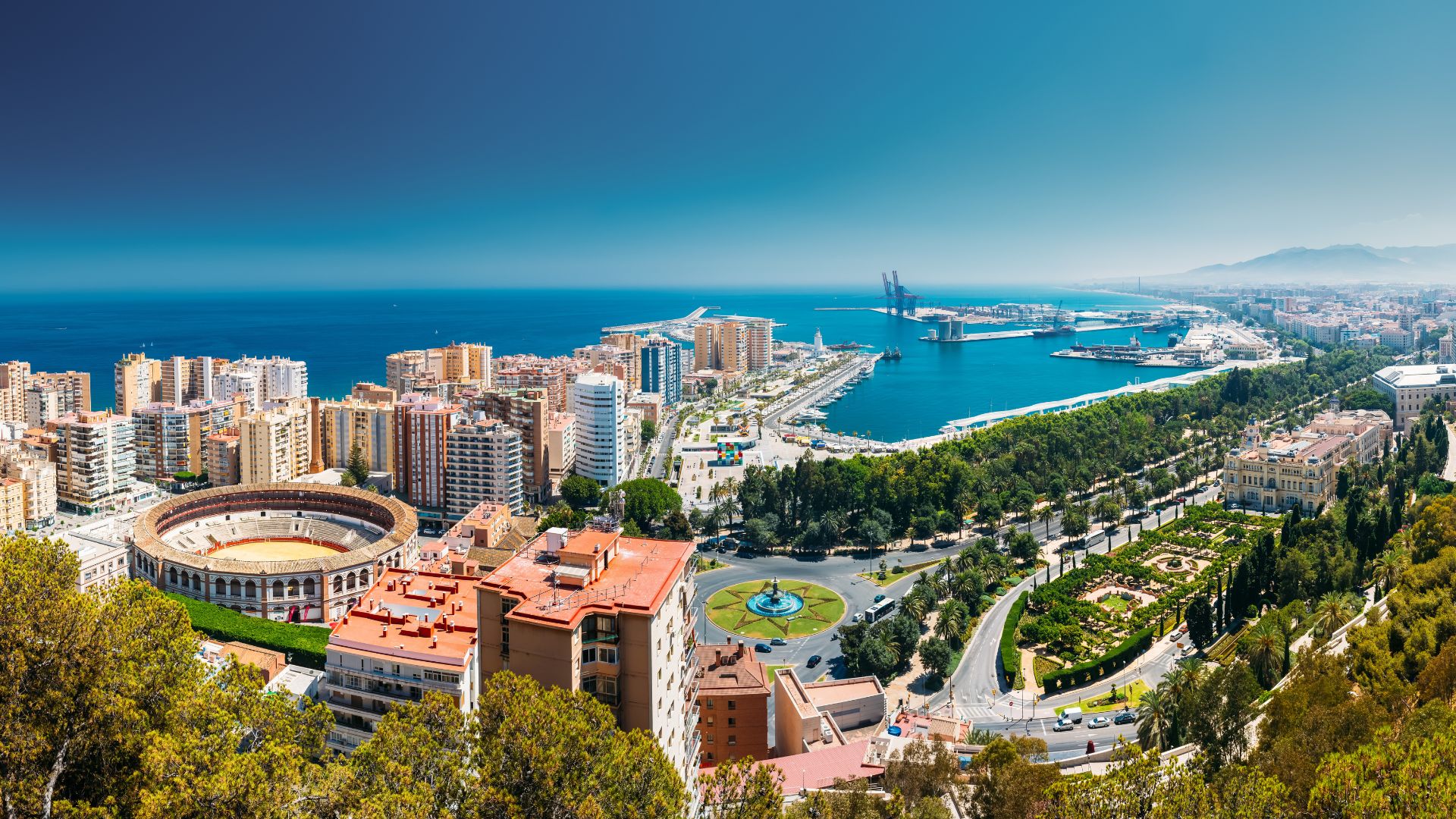 A panoramic aerial view of Málaga, Spain, showcasing a vibrant coastal city with a large bullring (Plaza de toros de La Malagueta) in the foreground, surrounded by urban buildings, lush green parks, and a bustling port leading out to the deep blue Mediterranean Sea under a clear sky. 