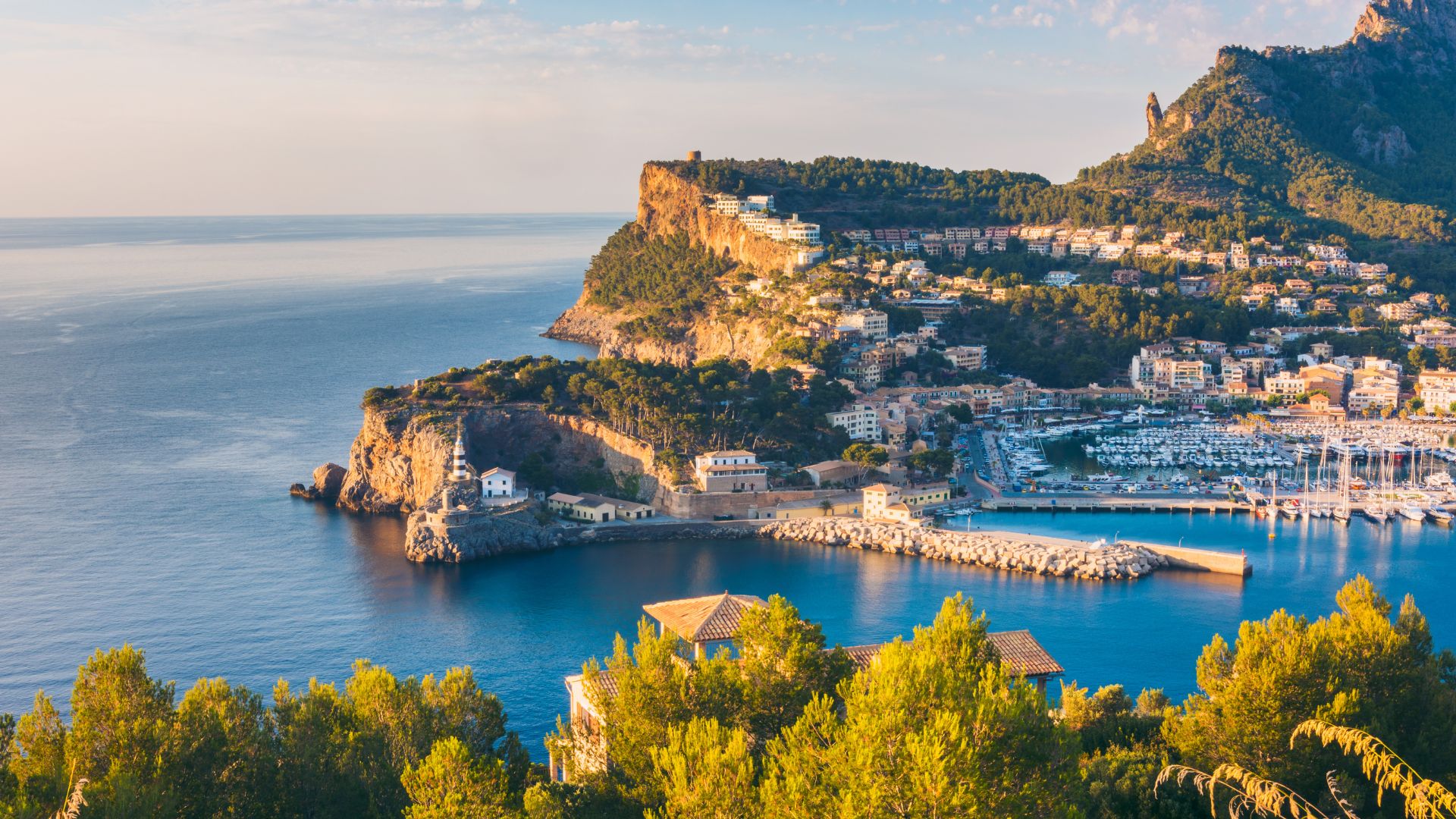 An aerial view of Port de Sóller in Mallorca, Spain, showing a scenic bay with a marina, coastal town, and surrounding mountainous landscape under a clear sky.