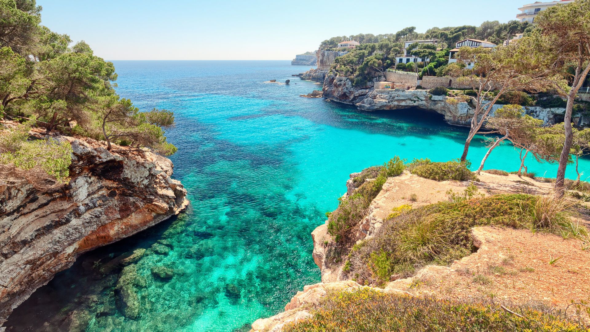 A scenic view of a turquoise cove in Mallorca, Spain, with clear water, rocky cliffs, and green foliage under a bright blue sky.