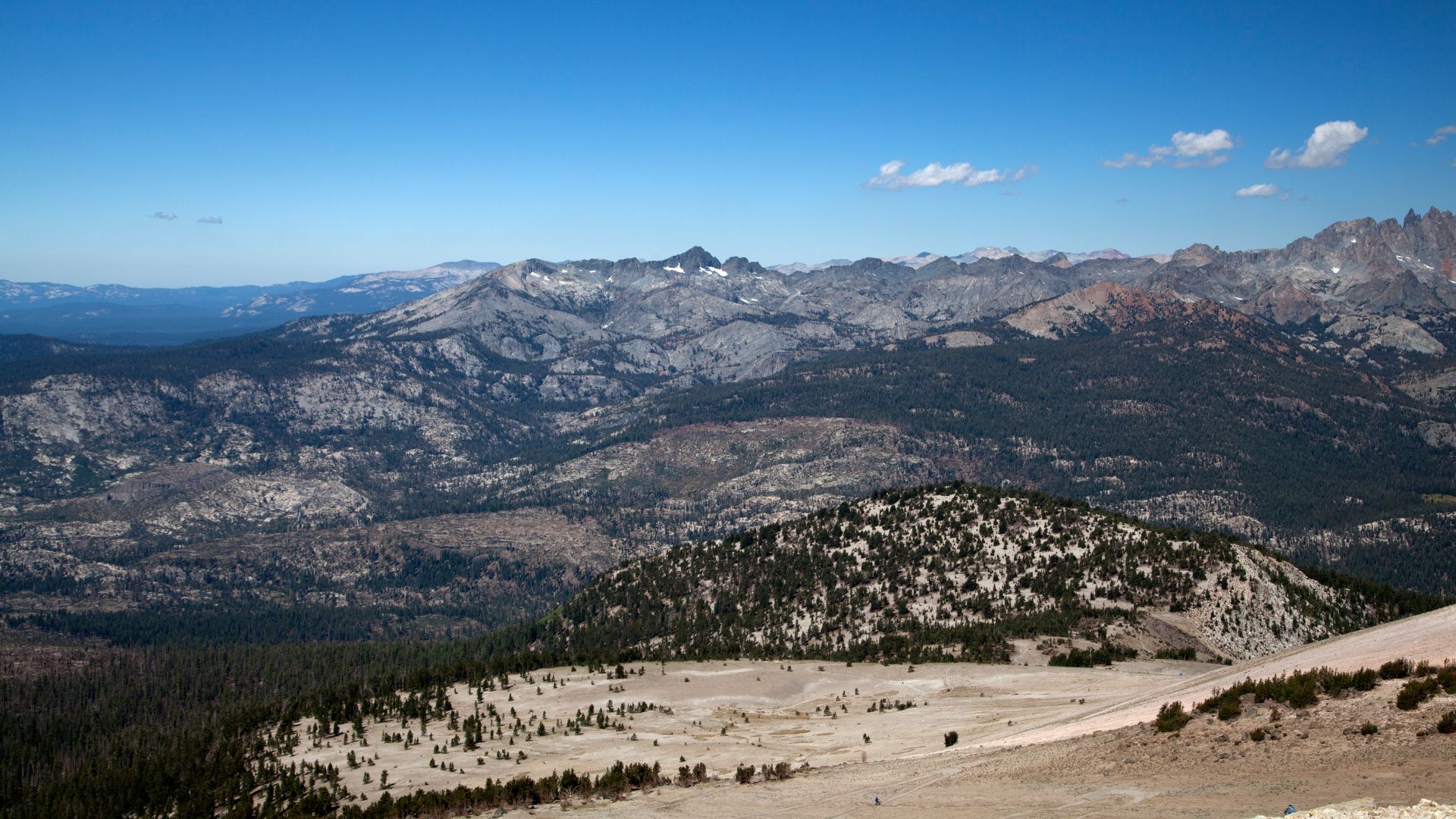 A wide shot of a mountainous landscape under a clear blue sky, showing forested hills in the foreground leading up to a range of rugged, snow-dusted peaks in the distance, with a sandy, sparsely vegetated slope in the immediate foreground.