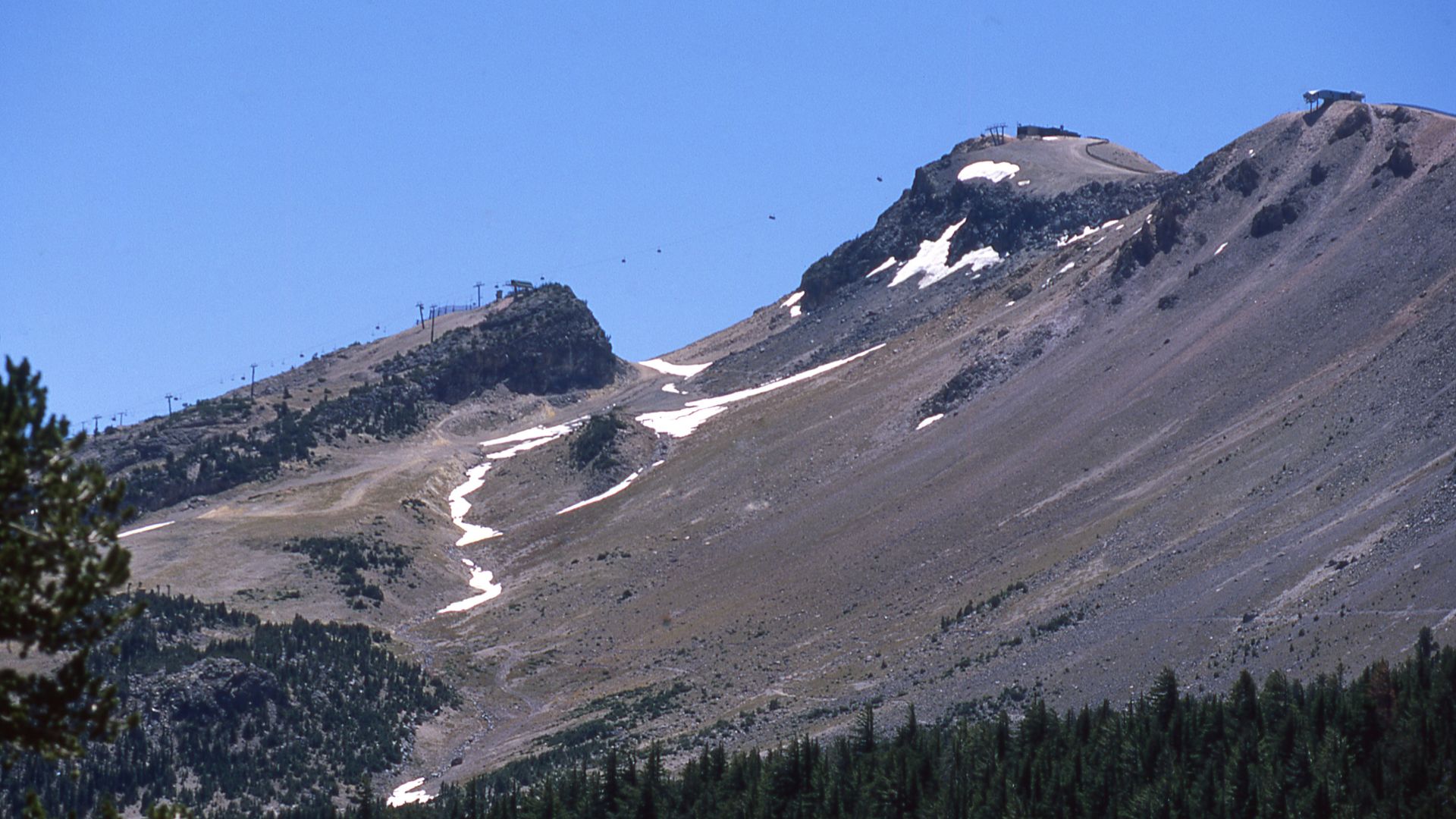 A wide shot of Mammoth Mountain under a clear blue sky, showing rocky slopes with patches of snow, several ski lifts, and a dense forest line at the base of the mountain.