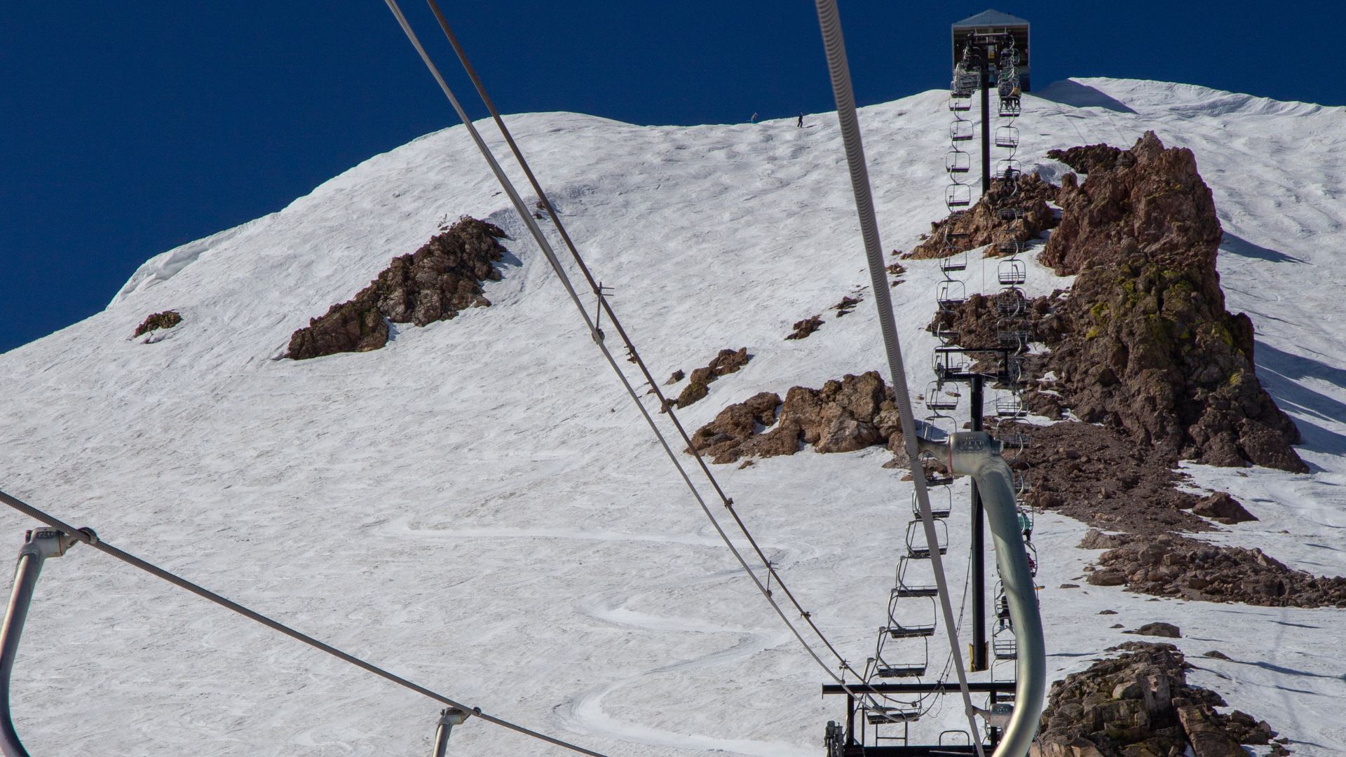 A snowy mountain slope with a ski lift ascending towards a summit structure under a clear blue sky.