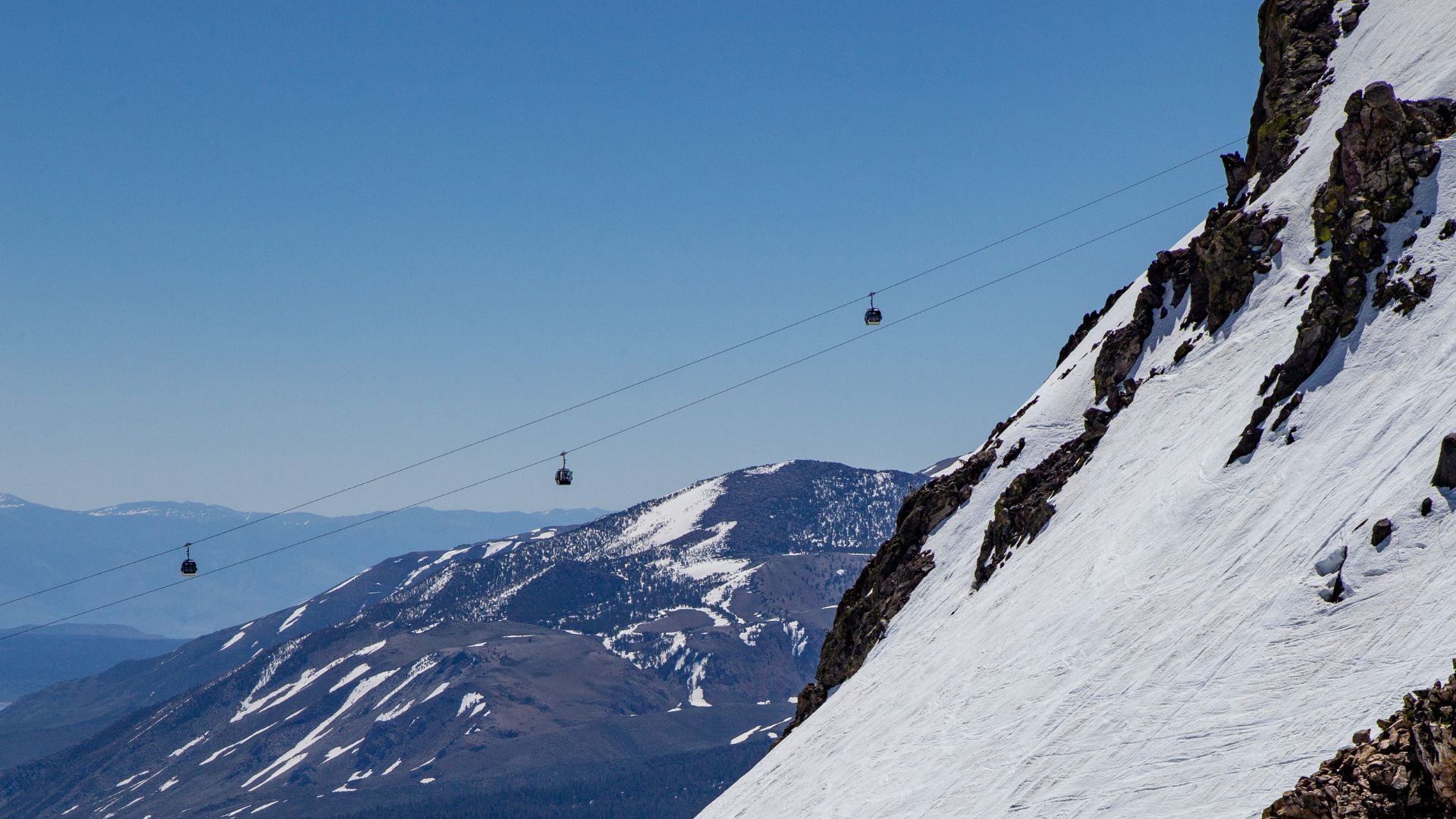 A clear blue sky stretches above a snow-covered mountain with a steep, rocky face on the right. In the distance, several gondola cars ascend and descend along cables, connecting to a distant, snow-capped mountain range under the same clear sky.