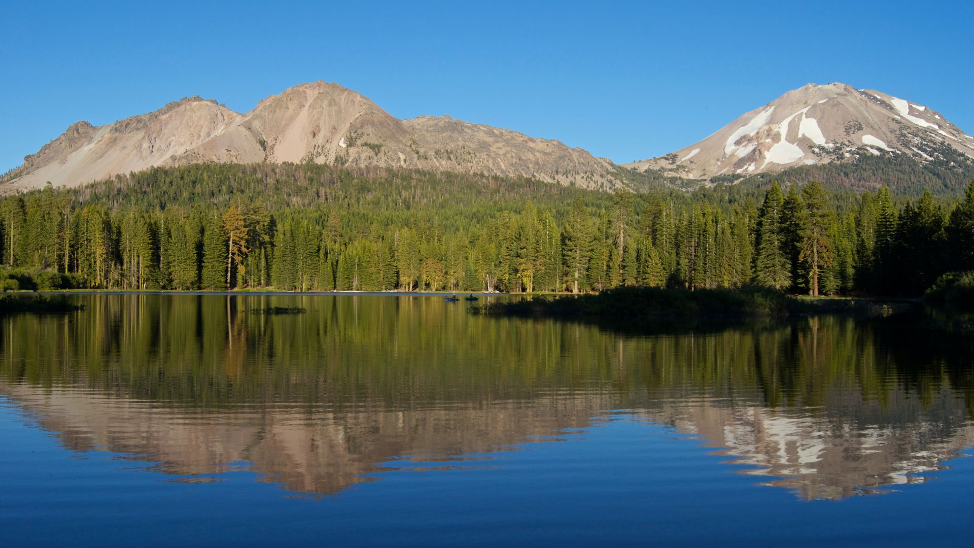 A panoramic view of a subalpine meadow with a stream winding through it, patches of snow in the foreground, and a dense evergreen forest leading up to a snow-capped volcanic peak under a clear blue sky.
