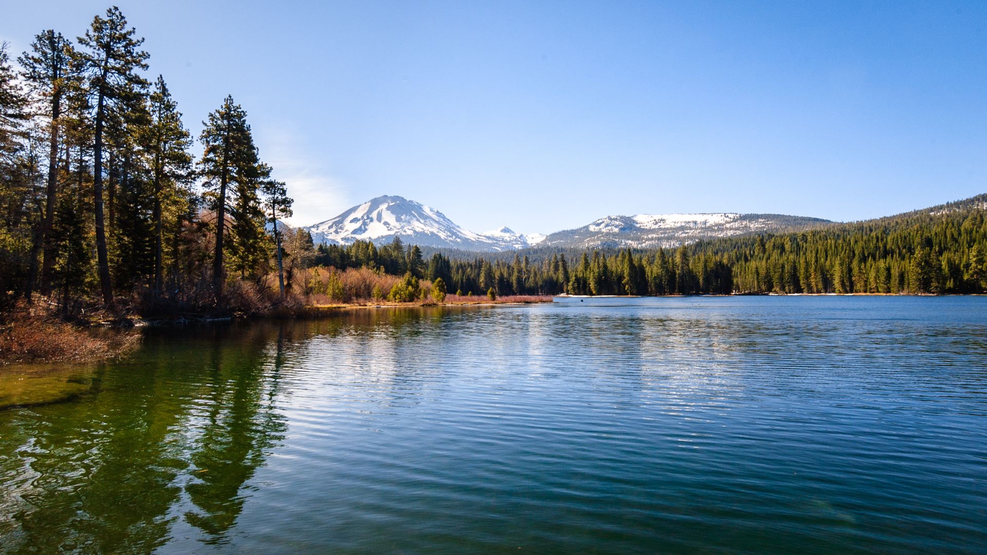 A serene lake, Manzanita Lake, reflects the surrounding evergreen forest and snow-capped peaks of Lassen Volcanic National Park under a clear blue sky.