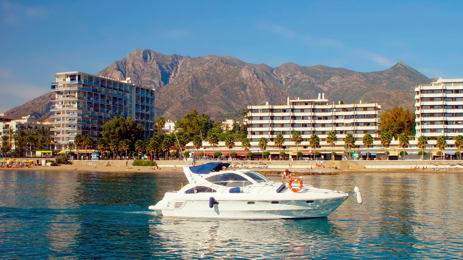 A white yacht cruises on calm blue water in a bay, with a bustling city featuring white buildings and palm trees lining a beach in the background, all set against a backdrop of large mountains under a clear blue sky.