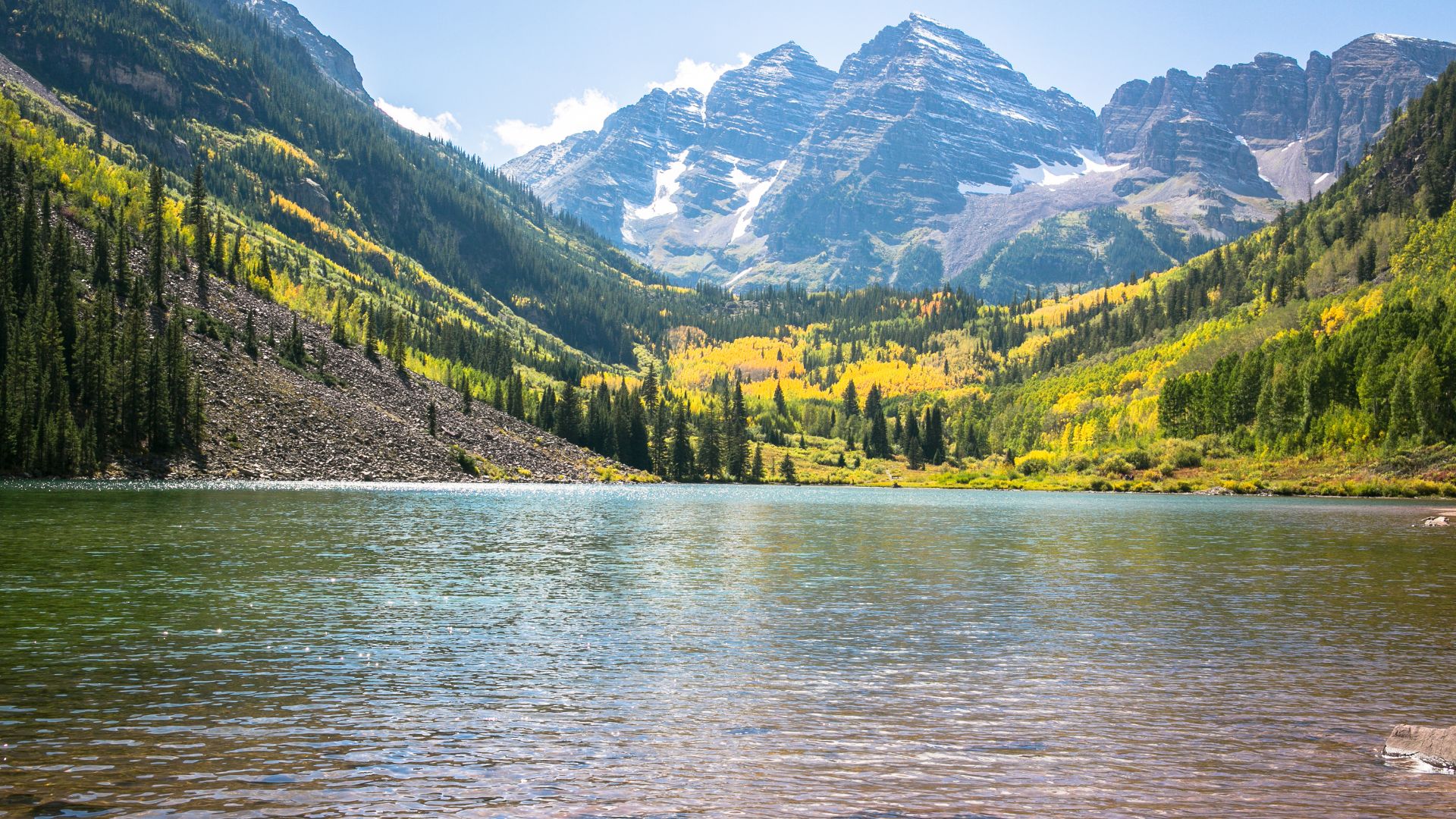 A scenic view of Maroon Lake reflecting the snow-capped Maroon Bells peaks, surrounded by vibrant golden aspen trees and green pine forests under a clear blue sky.