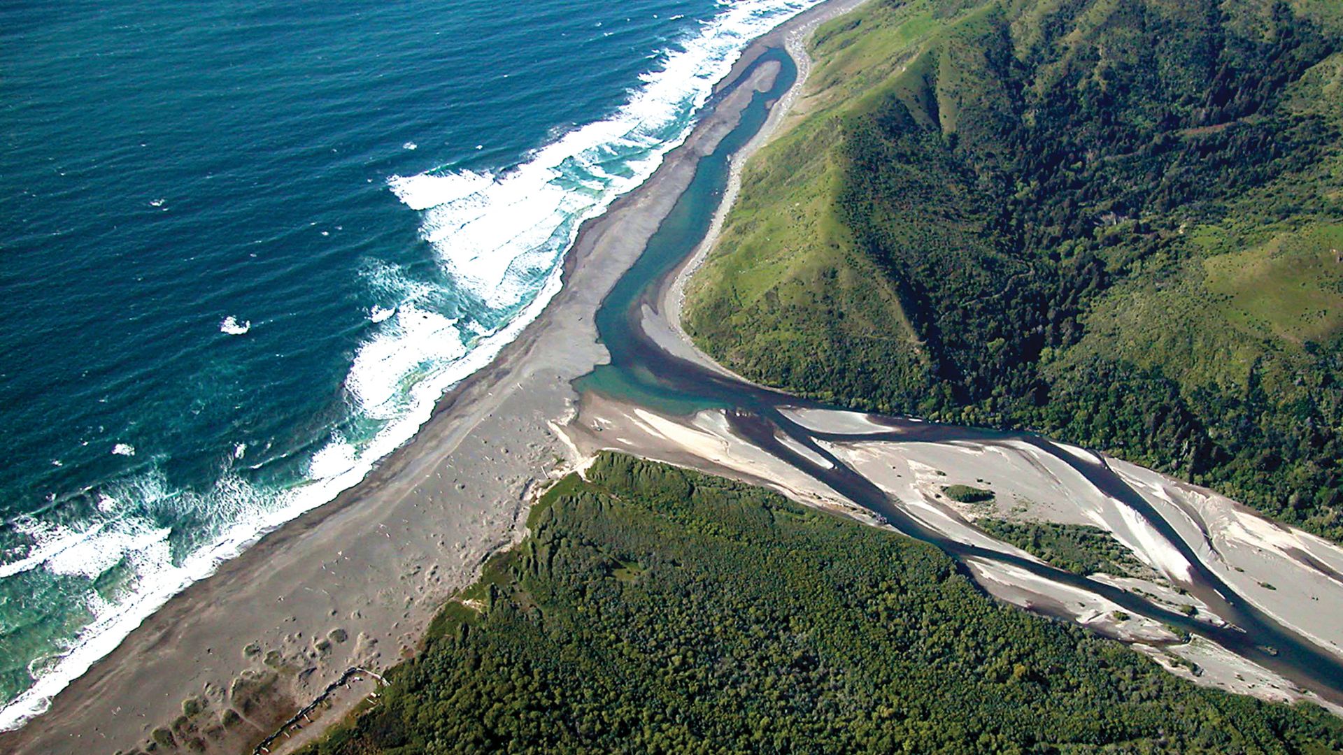 Aerial view of the Mattole River winding through green hills and emptying into the Pacific Ocean at a wide, dark sand beach, with waves crashing along the shoreline.