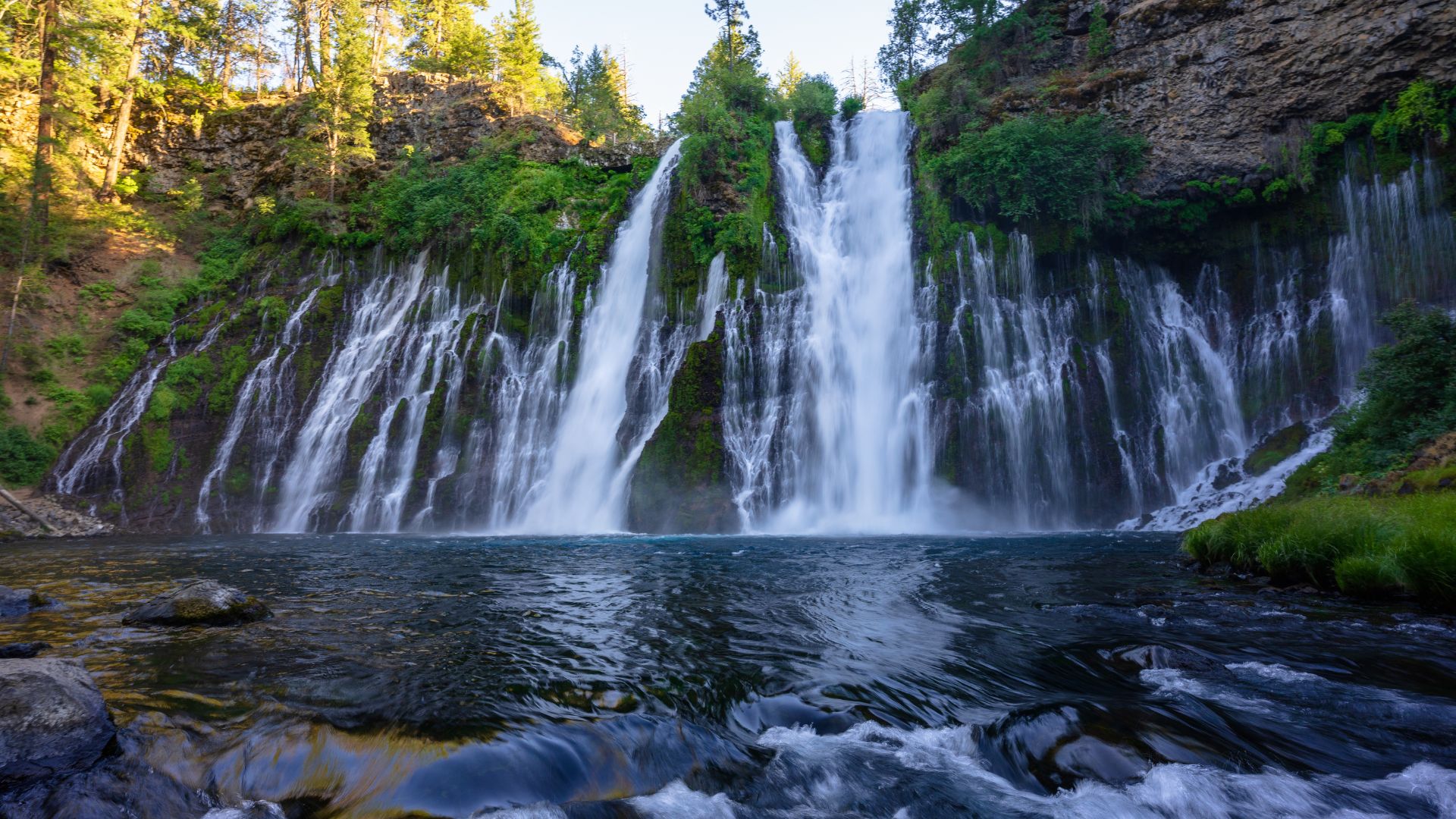 A wide shot of McArthur-Burney Falls, showcasing the 129-foot waterfall cascading into a pool below, surrounded by lush green foliage and trees under a clear sky.