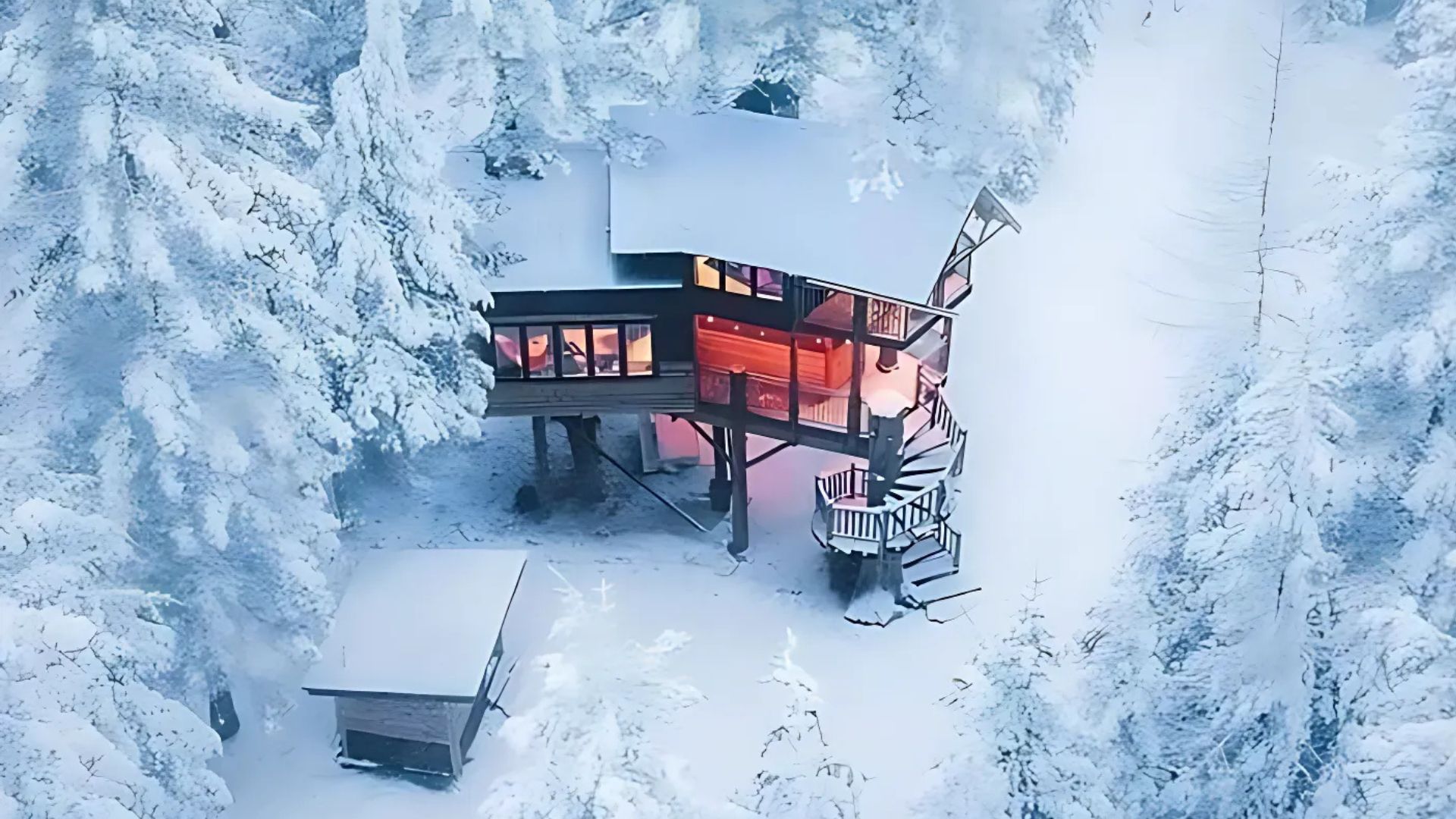 Aerial view of a rustic, multi-level treehouse with warm lights glowing from its windows, nestled among snow-covered evergreen trees in a winter forest landscape. A spiral staircase leads up to the main entrance, and a smaller structure is visible on the ground below.