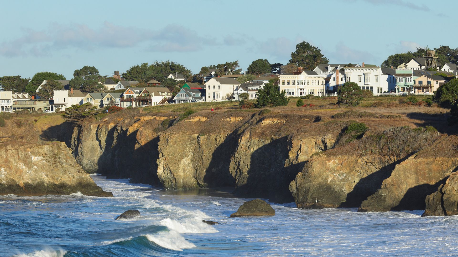 A picturesque coastal town perched on cliffs overlooking the Pacific Ocean, with houses and buildings visible on the bluff and waves crashing against the rocky shore below.