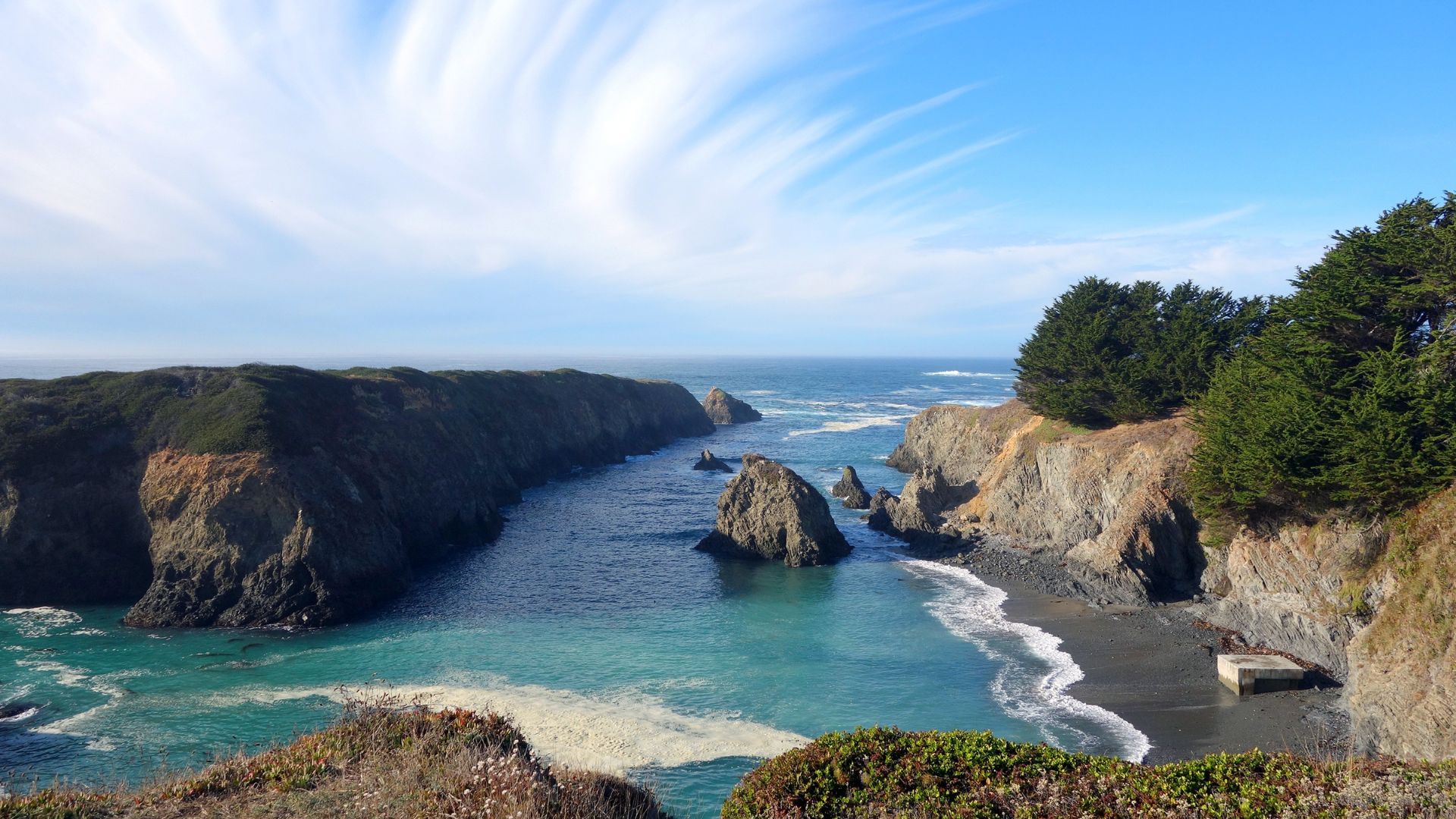 A wide shot of a sandy beach and a wooden trestle bridge along a rugged coastline under a clear blue sky, with scattered trees and distant bluffs.
