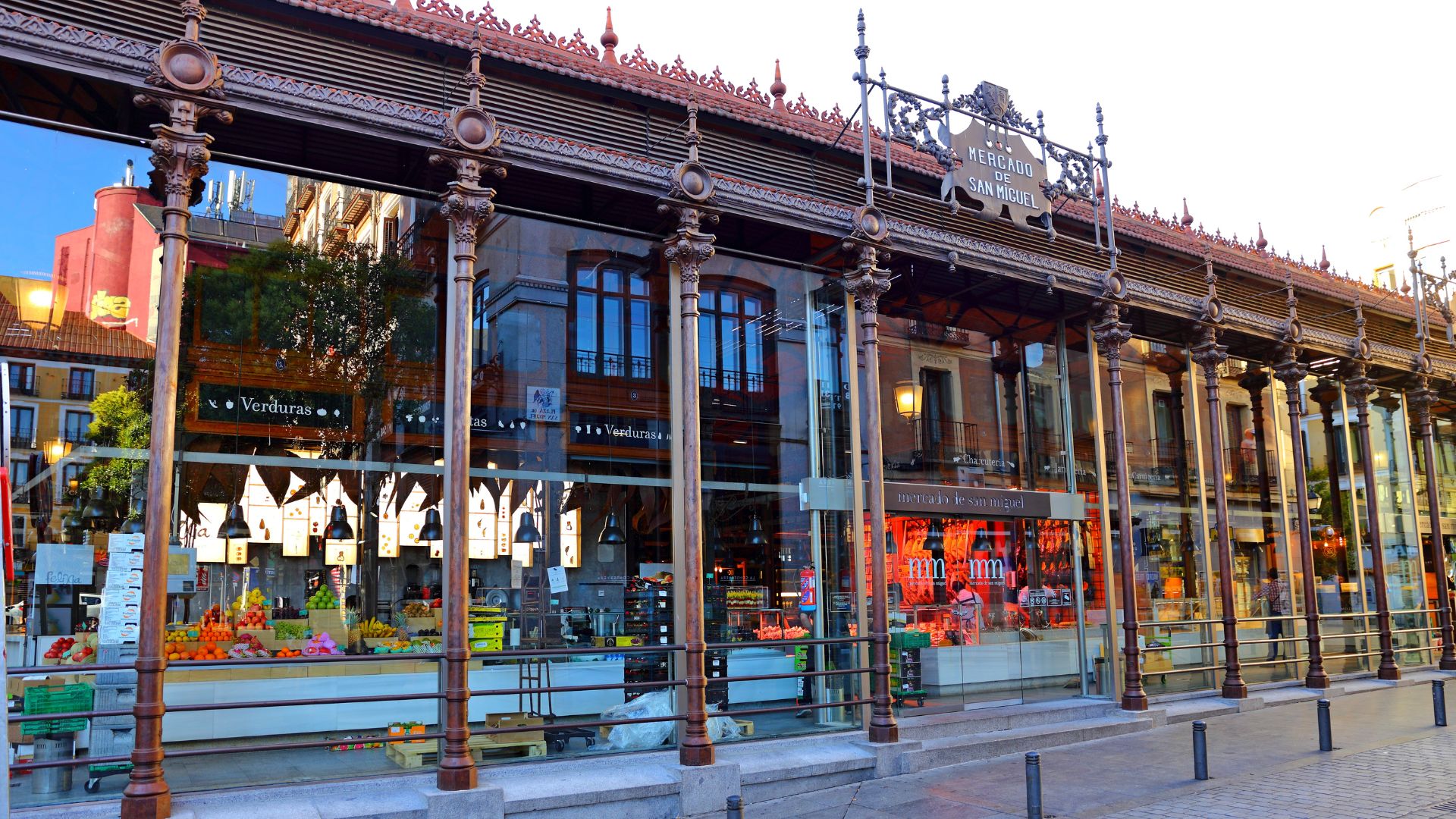A wide exterior shot of the Mercado de San Miguel in Madrid, Spain, a historic market building with a distinctive cast-iron and glass structure, showcasing its facade and the vibrant market stalls visible through the large glass windows.