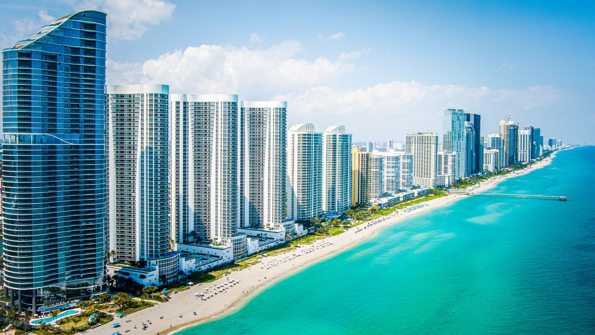 An aerial view of a vibrant coastline with a long stretch of sandy beach, turquoise ocean waters, and a dense array of modern high-rise buildings under a clear blue sky with scattered clouds.