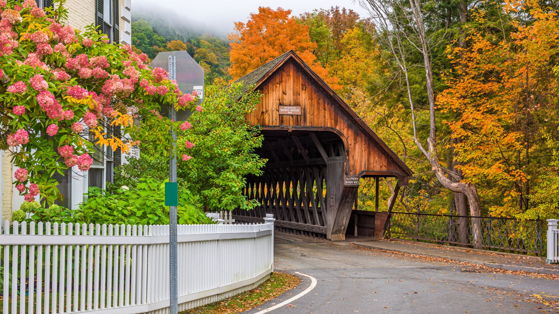 A wooden covered bridge spans a road and river, surrounded by vibrant autumn foliage and a white picket fence, with pink flowering bushes in the foreground.