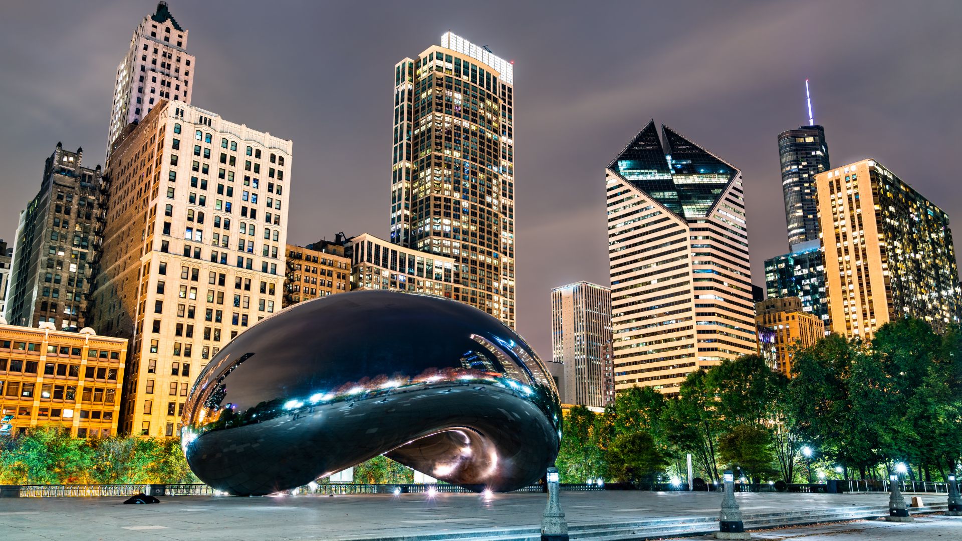 A nighttime view of Cloud Gate, a large, reflective bean-shaped sculpture, in Millennium Park, Chicago, with illuminated city skyscrapers in the background.