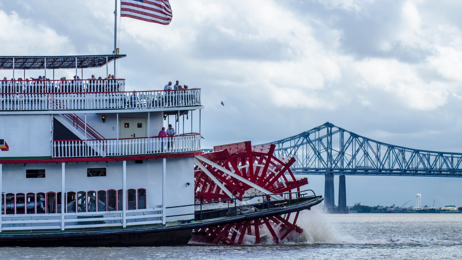 A large white paddlewheel steamboat with red paddles, the Steamboat Natchez, cruises on a river with a prominent bridge (Crescent City Connection) in the background under a cloudy sky.