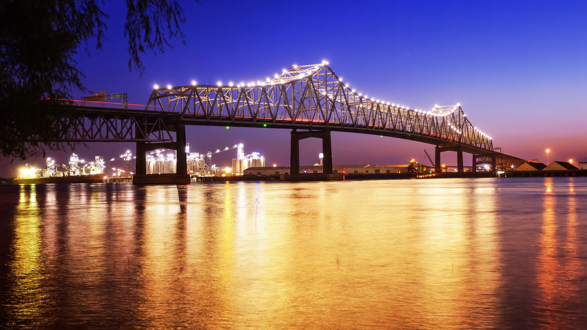 A nighttime view of the Horace Wilkinson Bridge (also known as the "New Bridge") in Baton Rouge, Louisiana, illuminated with lights, spanning across the Mississippi River, with reflections shimmering on the water and city lights visible in the background.