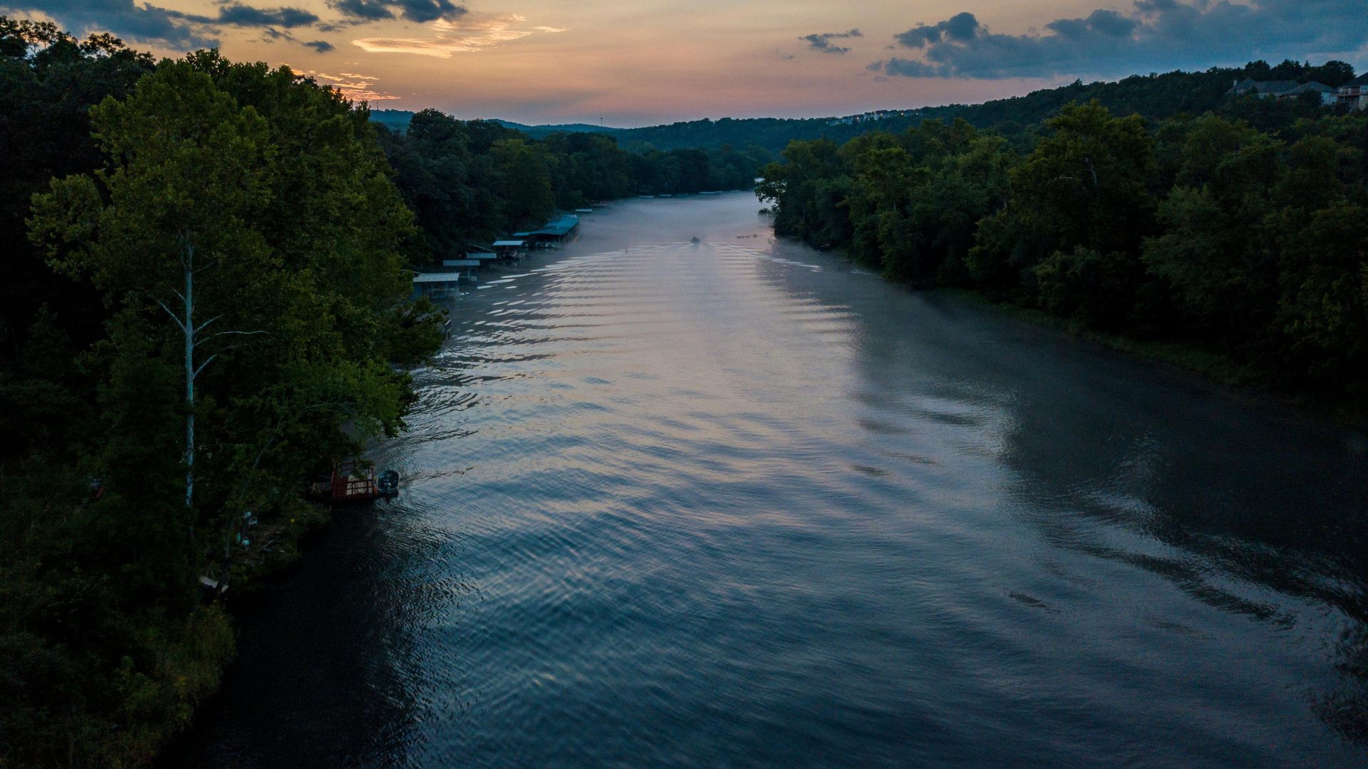 A wide river, likely the Missouri River, flows through a forested landscape under a dramatic twilight sky, with trees lining both banks and a few structures visible on the left bank in the distance.