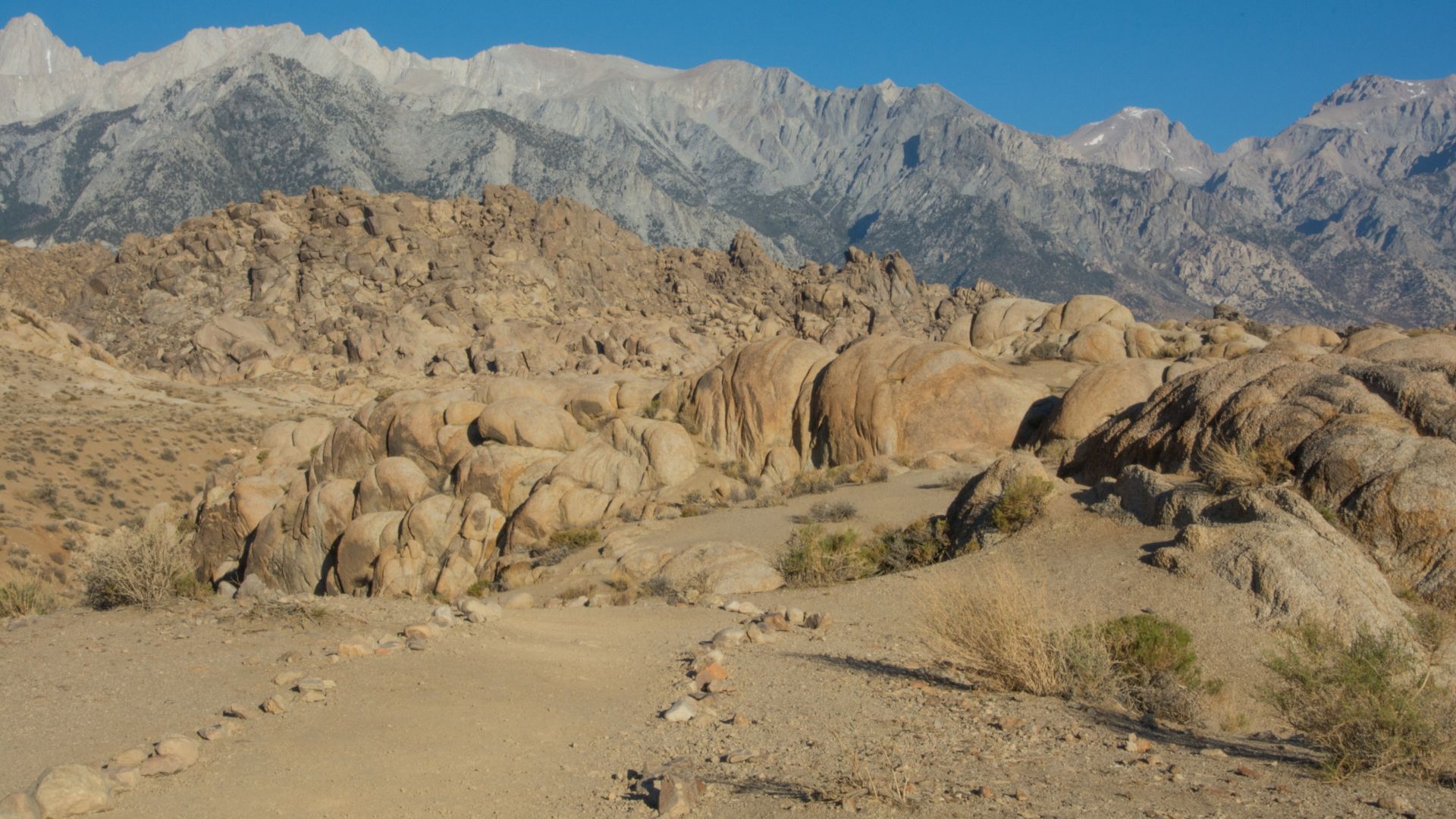A wide shot of a rocky, arid landscape with large, rounded rock formations in the foreground and middle ground, leading towards rugged, snow-capped mountains under a clear blue sky in the background.
