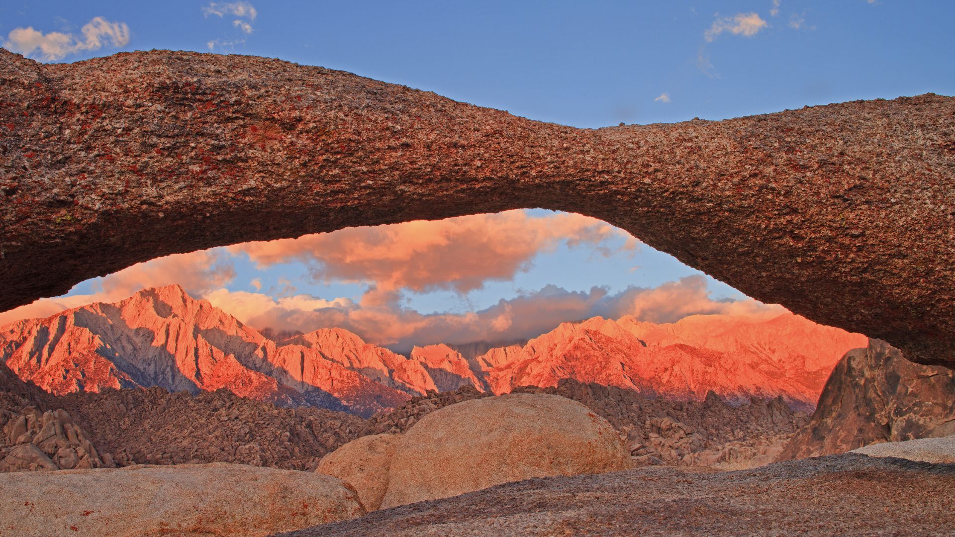 A natural rock arch, Mobius Arch, frames a vibrant sunset sky over the rugged, reddish peaks of the Sierra Nevada mountains in California's Alabama Hills.