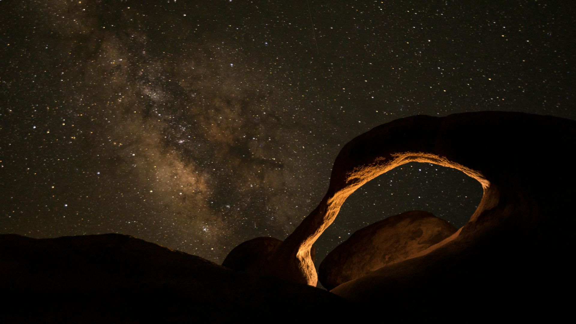 A natural stone arch, Mobius Arch, frames a view of distant mountains under a bright, sunlit sky with a prominent sunburst effect visible through the arch's opening. The arch is composed of light-colored rock, and the foreground shows rocky terrain.