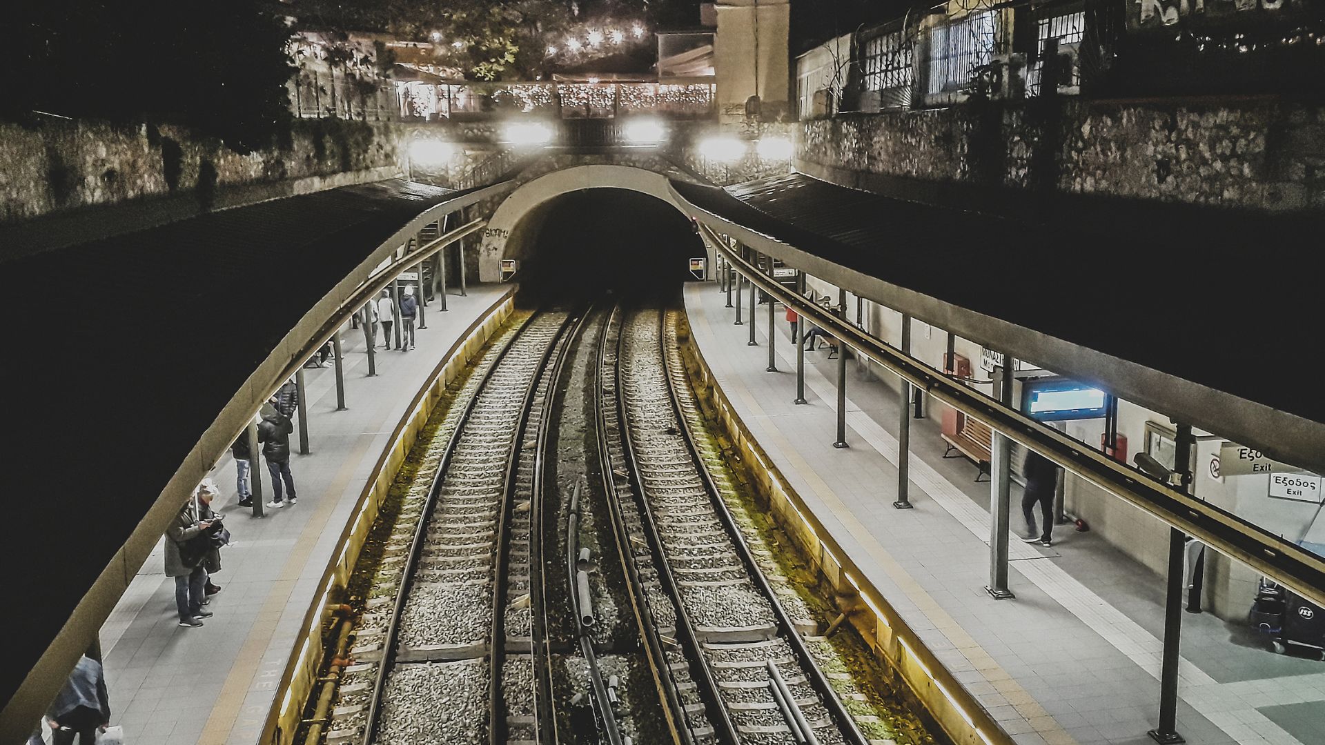 A nighttime, high-angle view of the Monastiraki Metro Station in Athens, Greece, showing train tracks leading into a tunnel, flanked by illuminated platforms with a few people visible.