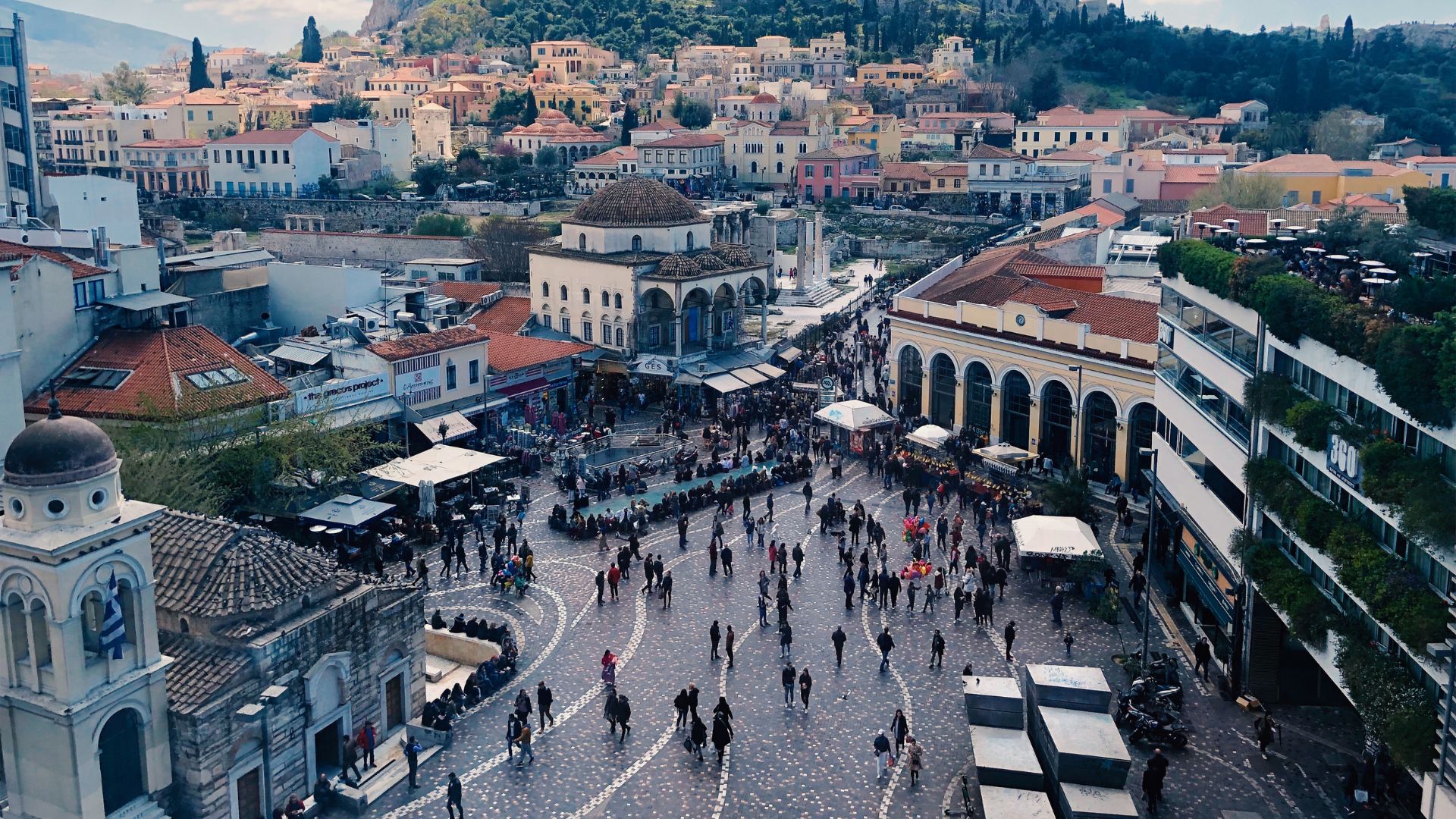 A bustling aerial view of Monastiraki Square in Athens, Greece, showing a large open plaza filled with people, surrounded by historic buildings and shops, with the Acropolis visible in the background.