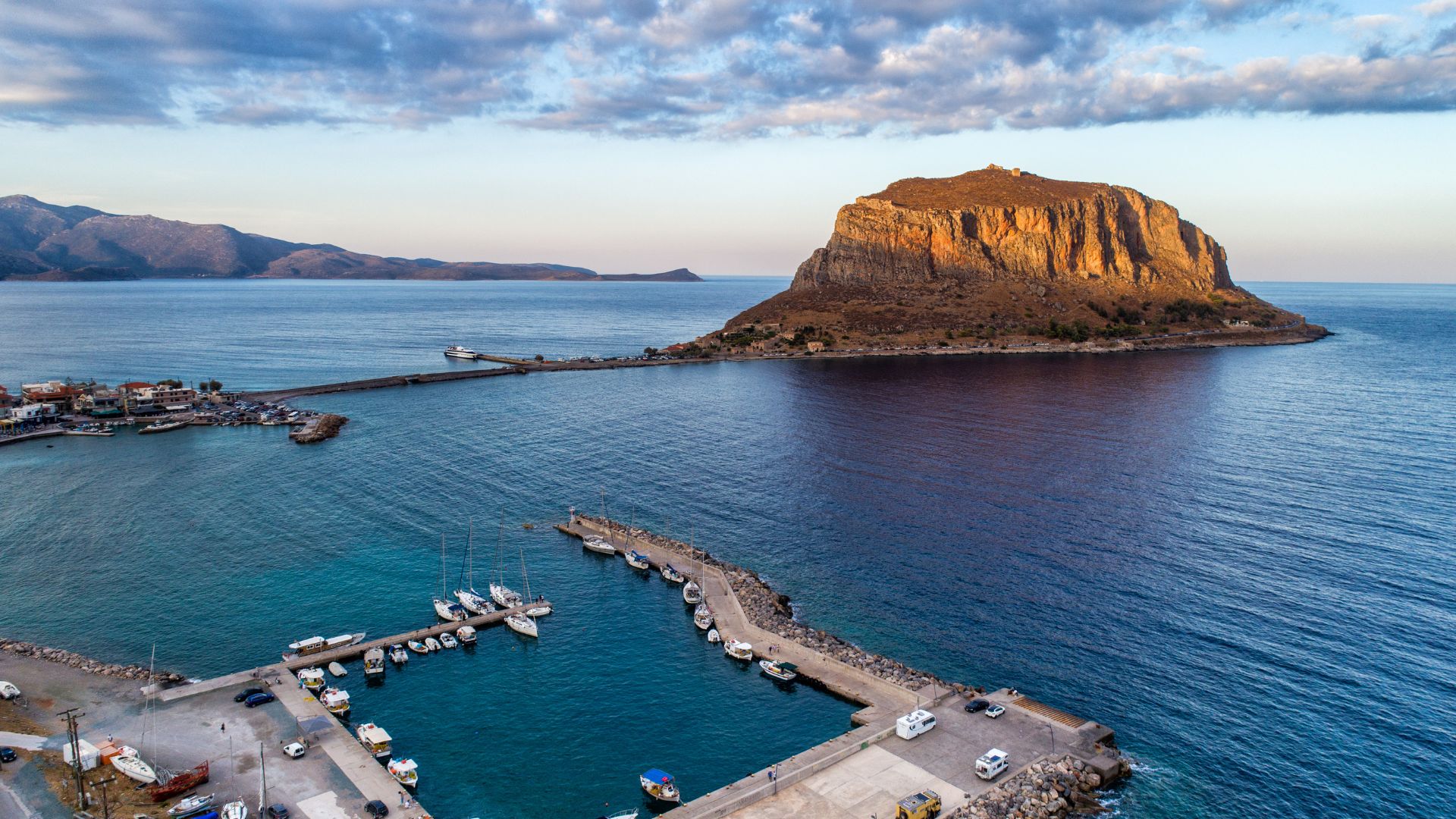 An aerial view of Monemvasia, Greece, showing a fortified medieval town built on a large rock formation connected to the mainland by a narrow causeway, with a harbor filled with boats in the foreground and calm blue waters extending to the horizon under a partly cloudy sky.