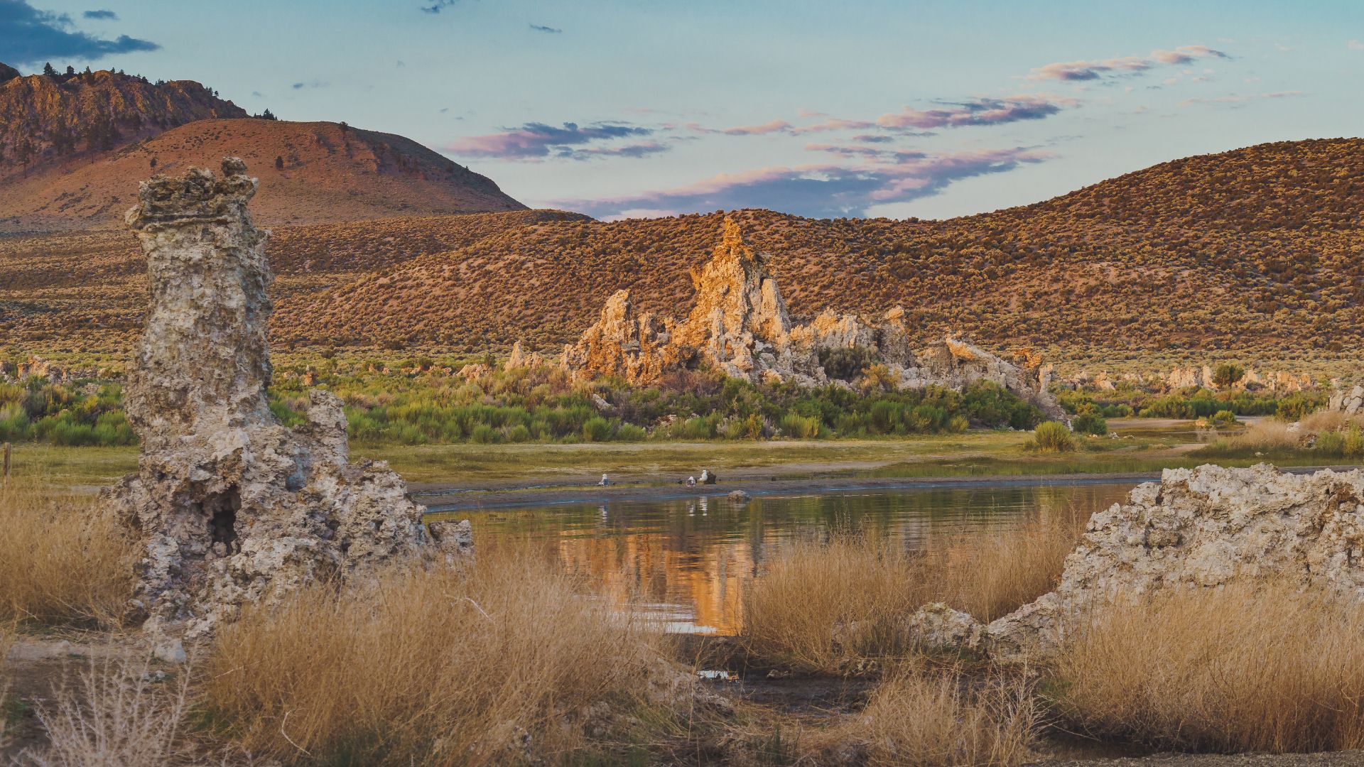 A wide-angle shot of Mono Lake in California at dusk, showcasing the unique limestone tufa towers rising from the water, with dry grasses in the foreground and mountains under a colorful sky in the background. 