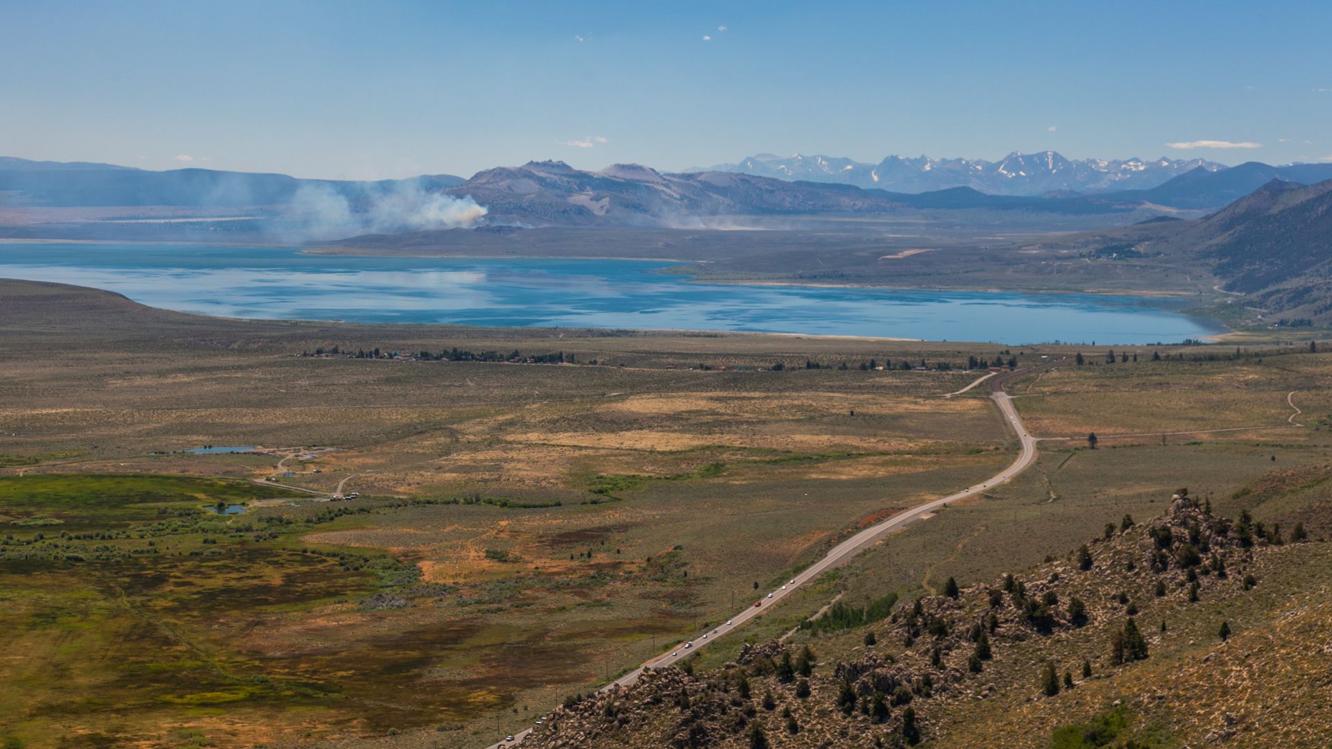 A wide aerial view of Mono Lake, a large blue lake with a mountainous backdrop, surrounded by a vast, dry landscape with a winding road leading towards the lake. Haze or smoke can be seen rising from the far side of the lake.