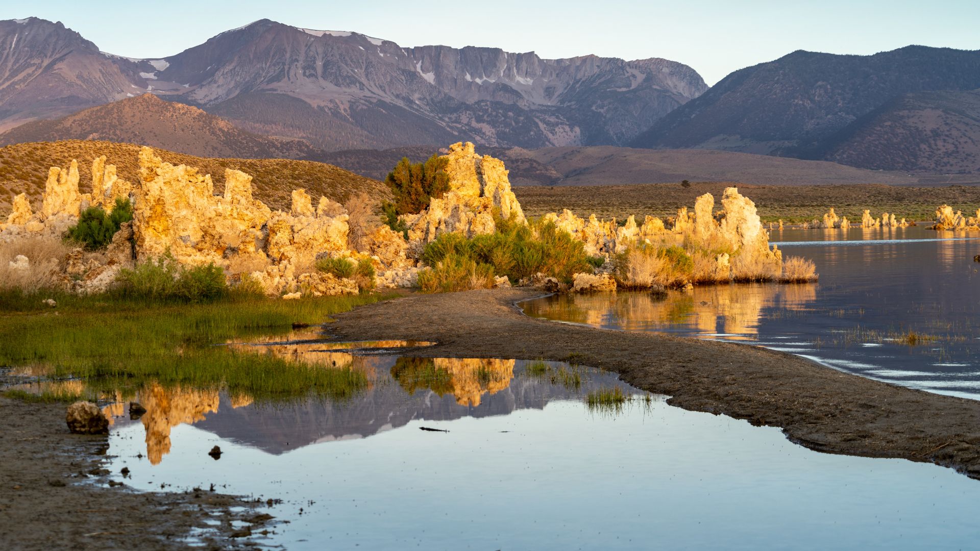 A serene landscape photo of Mono Lake at sunset, featuring prominent, light-colored tufa towers rising from the water, with reflections of the formations and distant mountains visible in the calm lake surface.