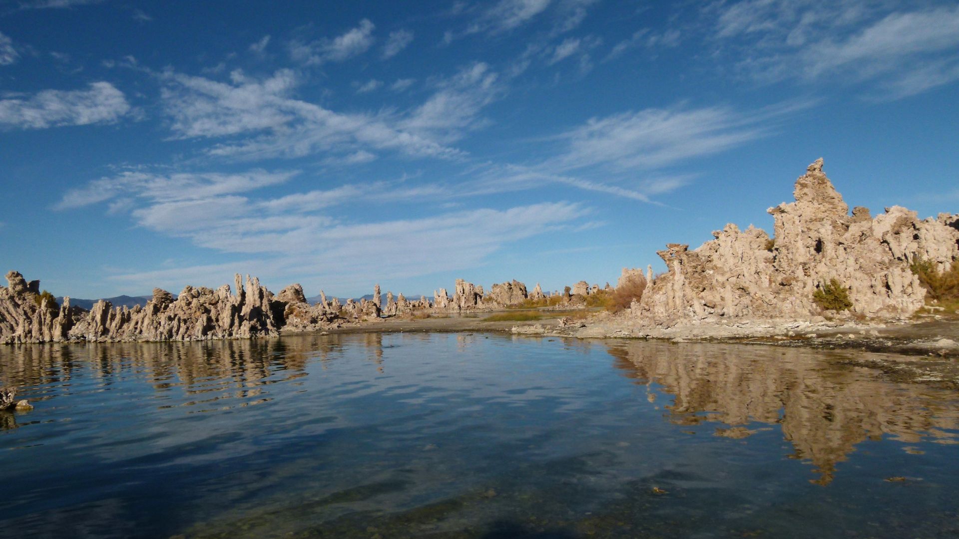 A panoramic view of Mono Lake in California under a clear blue sky with scattered clouds, featuring numerous light-colored tufa towers rising from the water and along the shoreline, reflected in the calm lake surface.