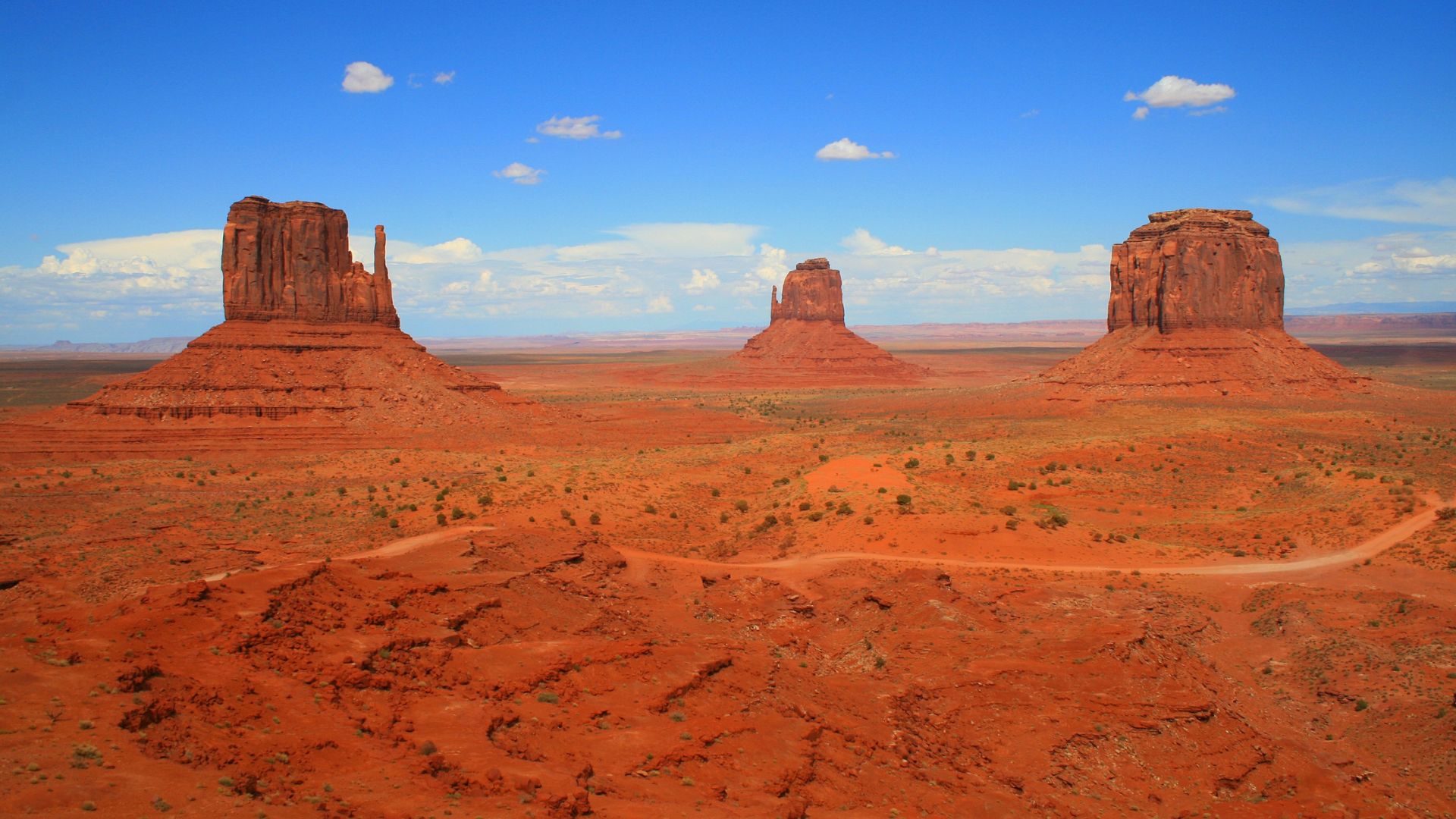 A wide shot of Monument Valley under a clear blue sky with scattered clouds, showcasing three prominent sandstone buttes rising from the vast, reddish-orange desert landscape, with a dirt road winding through the foreground.