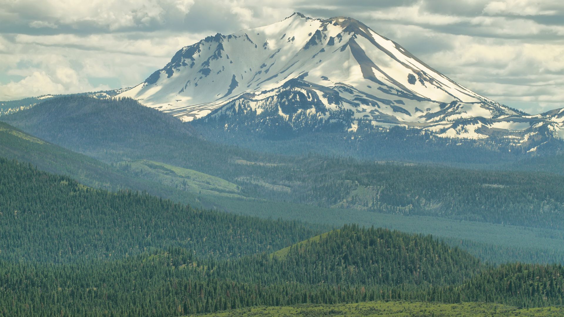 A snow-capped Mount Lassen rises above a dense, green forest under a cloudy sky in California.